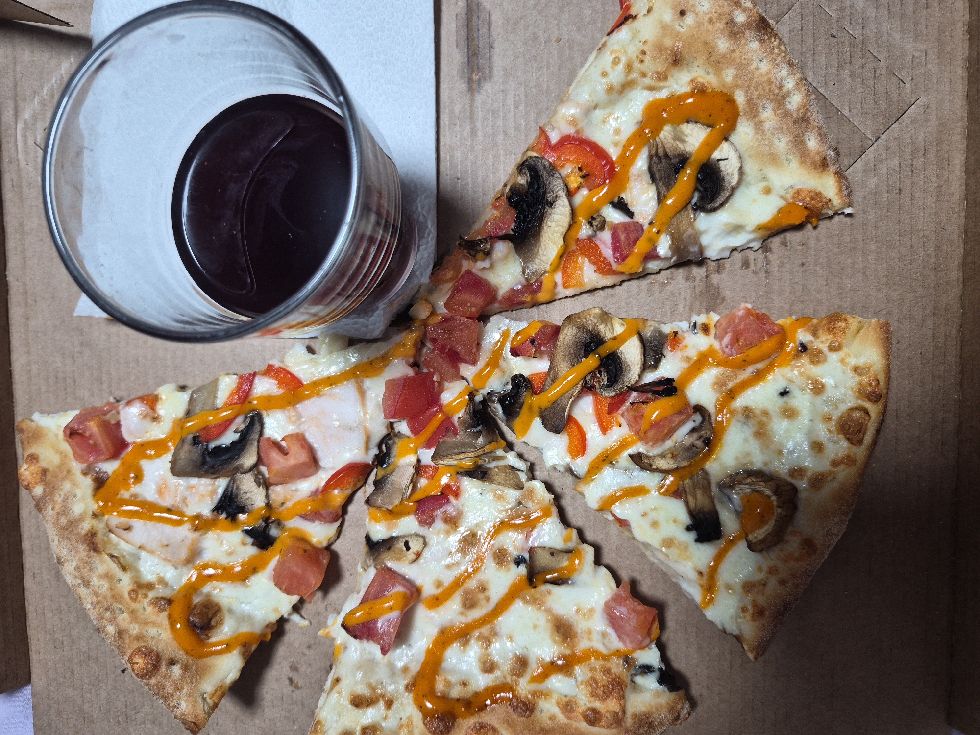 A medium shot, taken from above, shows four slices of pizza and a glass of dark liquid. The pizza is in a cardboard pizza box. The crust is golden brown, and the toppings include cheese, tomatoes, mushrooms, and red peppers. An orange sauce is drizzled over the pizza. The glass is clear and cylindrical, containing a dark red liquid, possibly wine or juice. The glass rests on a white paper napkin. The background is a brown cardboard pizza box with some textured lines. The location is Unknown, Unknown.