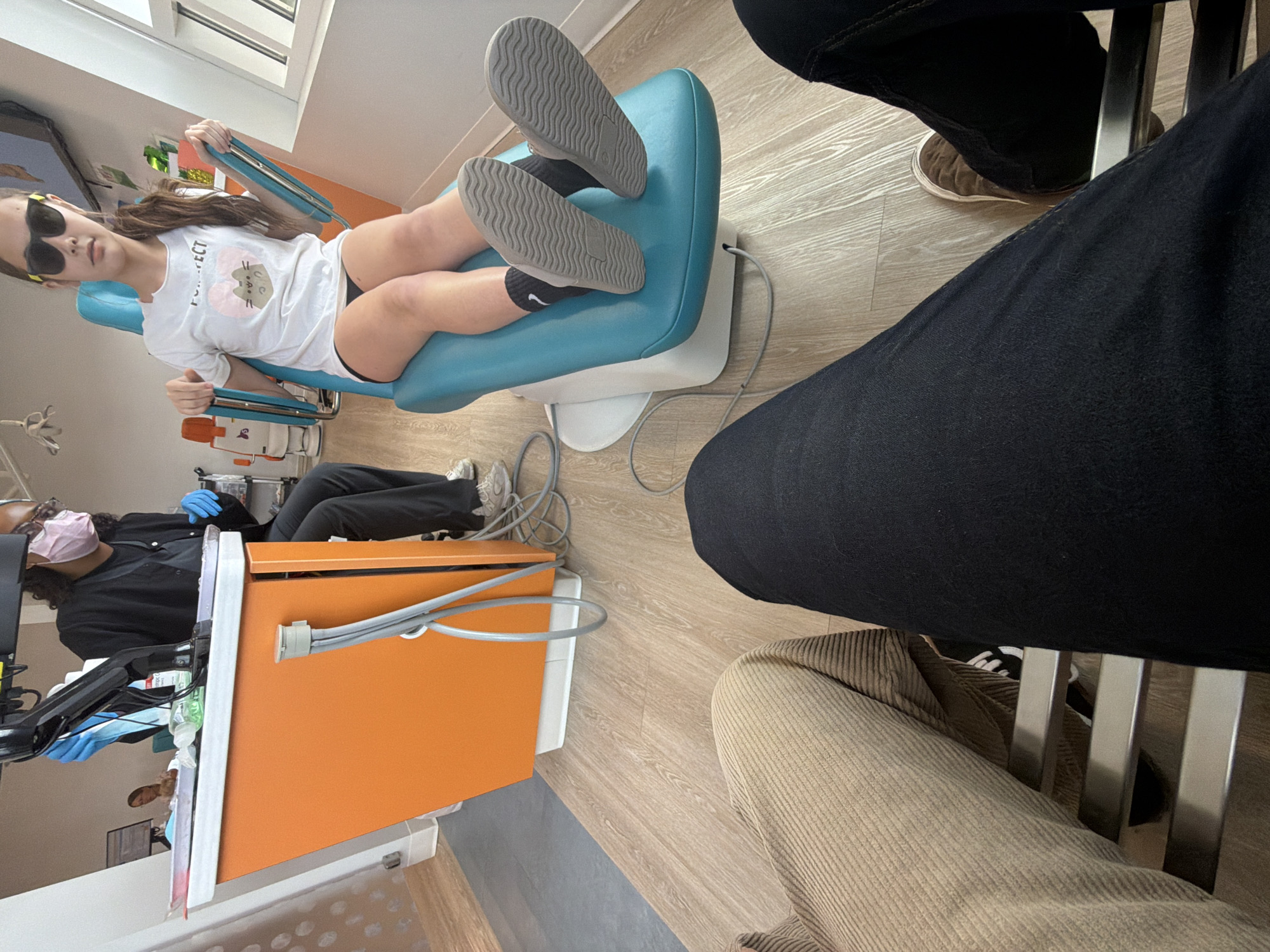 In a brightly lit dental office, a young girl wearing sunglasses sits in a teal dental chair, her legs crossed. She is wearing a white t-shirt with a cartoon cat graphic and black shorts. A dental professional in dark scrubs and a light pink mask stands behind her, wearing blue gloves and holding medical equipment. The office features a prominent orange dental cart with attached tubing. In the foreground, the legs of two individuals are visible, one wearing tan corduroy pants and the other in dark denim jeans. The floor is made of light-colored wood laminate. The overall impression is a sterile yet modern dental environment.