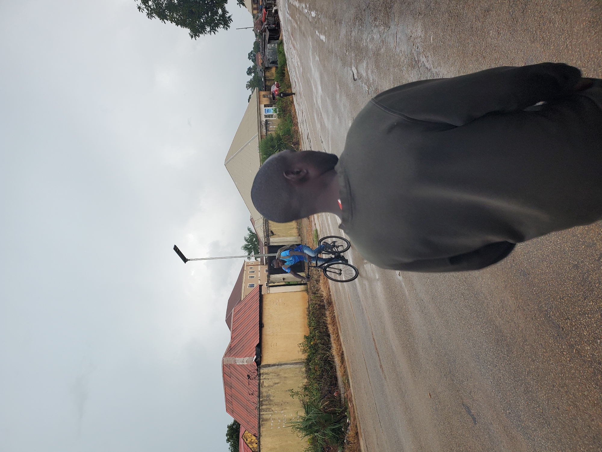 The media file depicts a street scene in Barikin Gidar Kanat, Nigeria. The primary subjects are two individuals. One is in the foreground, facing away from the camera, with a dark-colored hoodie. The other individual is further down the street, on a bicycle. This person is wearing a blue t-shirt and jeans and appears to be riding the bicycle. The scene is a paved road, which appears wet, suggesting recent rain or damp conditions. The time of day is unclear, but the overcast sky hints at potentially gloomy or rainy weather.
In the background, there are buildings with red and yellow exteriors, typical of the area. There are also some trees visible, including a large one at the top left of the frame. A street lamp is also present, with a distinctive solar panel attachment. The overall impression is that of a typical day in a residential or mixed-use area in Nigeria, with the focus on the street and the people present. There is no visible text in the image. The emotional tone is neutral, documenting a moment in time.