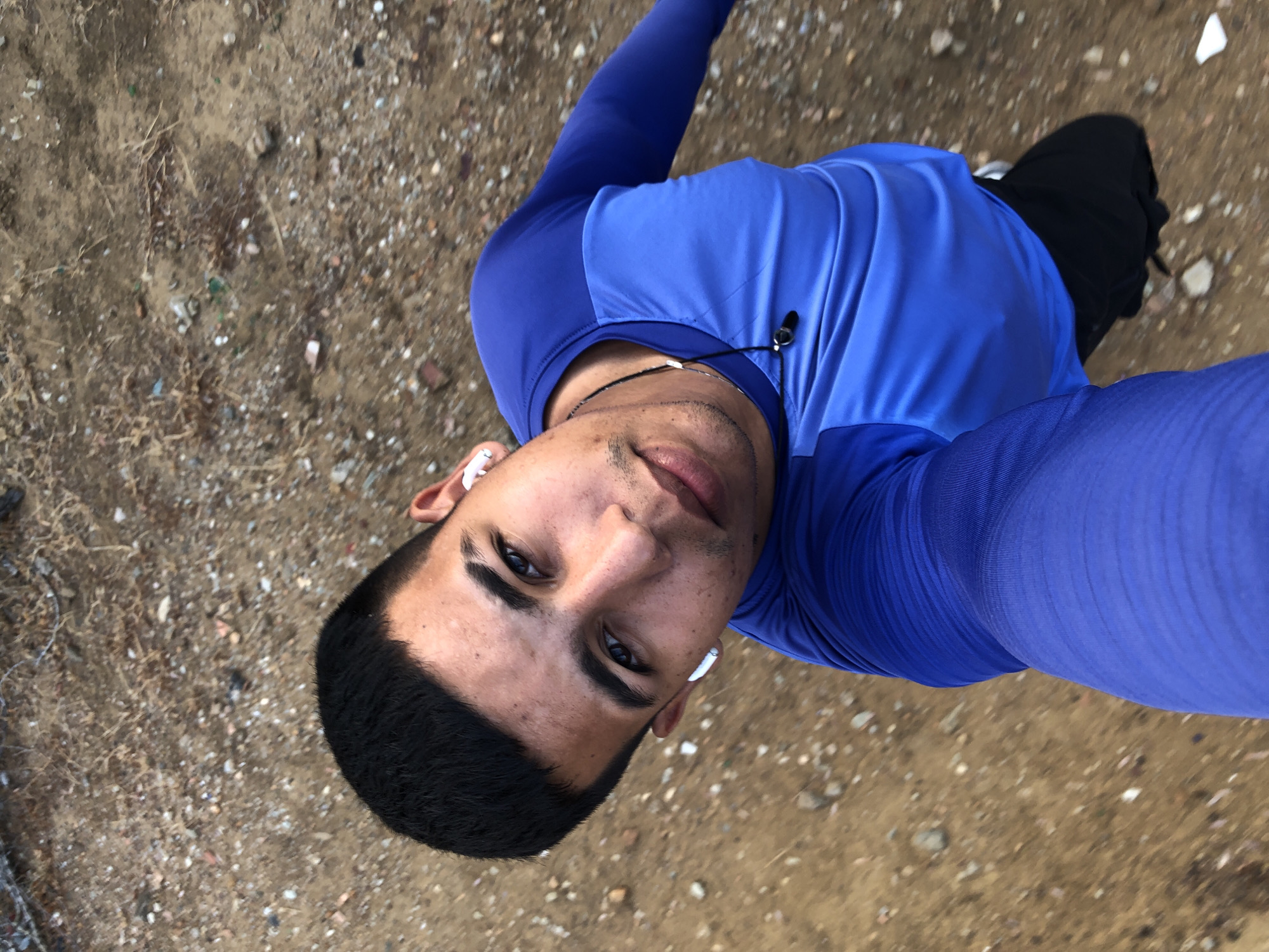 This high-angle selfie captures a young man looking directly into the camera from an overhead perspective. He is the central subject, positioned diagonally across the frame. He has dark, neatly trimmed hair, dark eyes, and slight facial stubble. His expression is neutral and direct.
The man is wearing an athletic, long-sleeved shirt that features two shades of blue: the sleeves are a darker royal blue, and the torso is a lighter, brighter blue. He has white wireless earbuds in both ears and a black wired earphone or microphone with a clip hanging around his neck. Part of what appears to be black shorts or pants is visible at the bottom right of the image.
The background is an outdoor, unpaved ground surface, consisting of dirt and gravel, with numerous small stones, pebbles, and some dry vegetation or debris scattered throughout. The texture suggests a rough, natural terrain, possibly a trail, undeveloped area, or dirt road. The lighting indicates daytime conditions, with diffused natural light illuminating the scene.
The image depicts the man taking a self-portrait outdoors, possibly during physical activity such as walking or exercising. There is no visible text in the image. The setting, characterized by dry, earthy ground, is consistent with outdoor environments found in regions like Altagracia, Venezuela.
