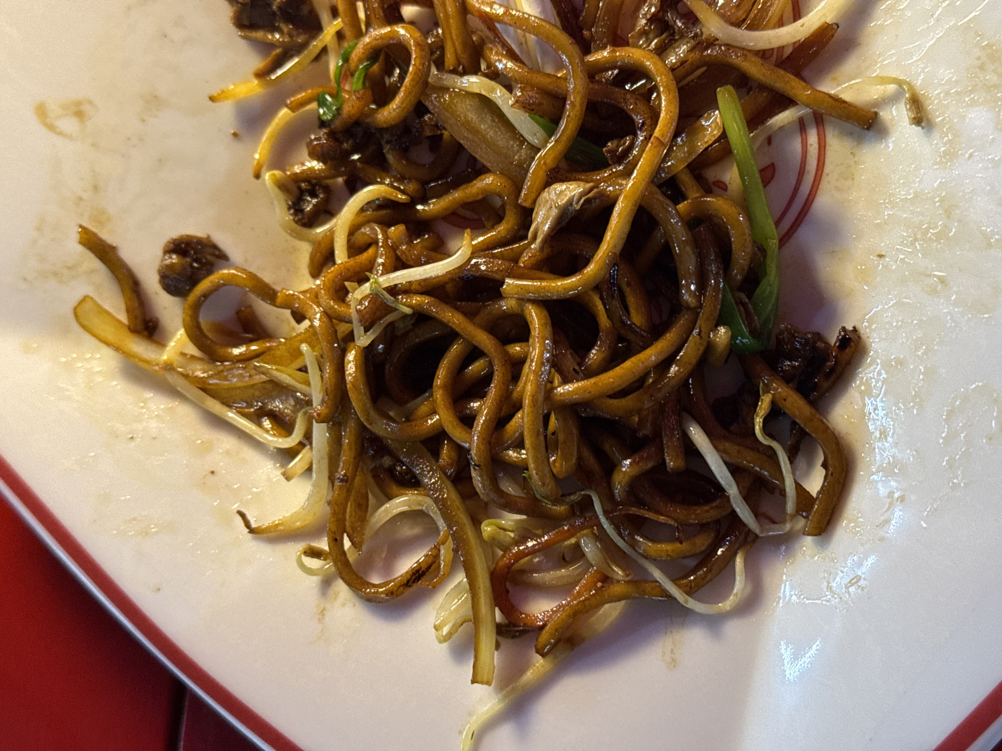 A close-up shot presents a plate of mie goreng, an Indonesian fried noodle dish, likely photographed in Munggu, Indonesia. The noodles are dark brown, glossy with sauce, and intermixed with bean sprouts and green onions. The noodles occupy the majority of the frame, arranged somewhat haphazardly on a white plate rimmed with red. The plate appears to be slightly dirty with residual food stains.