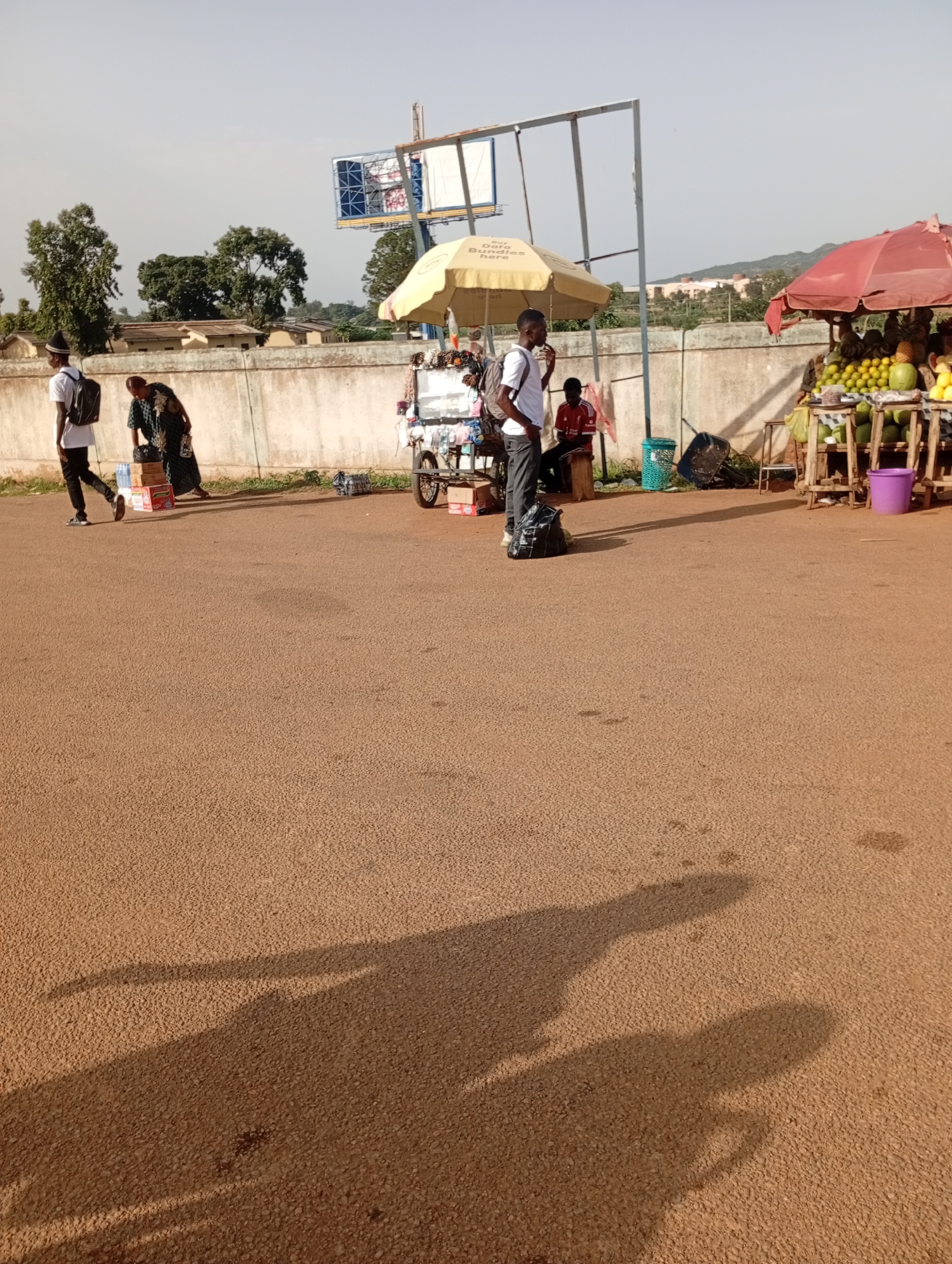 The image depicts an outdoor scene, likely a roadside market in Rafin Par, Nigeria. In the foreground, a sandy-colored paved surface fills the bottom half of the frame, with distinct shadows cast by individuals and structures, suggesting a bright, sunny day.
In the middle ground, several people are visible. To the left, a young man wearing a white t-shirt, black pants, and a black hat walks to the left, carrying a black backpack. Beside him, another person, appearing to be a woman, is bending down, dressed in a dark patterned garment, and has several boxes stacked near her. Further to the right, a young man in a white t-shirt and dark pants stands near a yellow umbrella, also carrying a backpack. Behind him, a person in a red shirt is seated on a stool.
Various objects are present, including multiple stacked boxes, some appearing to be food items. A small cart with a yellow umbrella is visible, containing what looks like packaged goods or snacks. To the far right, a stall under a red umbrella displays a variety of fruits, including oranges and what appear to be coconuts or similar round fruits. A purple bucket is placed near the fruit stall. Several green plastic baskets and a wheelbarrow are also scattered in the area.
In the background, a low concrete wall runs horizontally across the image, with some vegetation growing along its base. Beyond the wall, buildings and trees are visible, with a slight hill in the distance. A large billboard structure with blue and white panels is positioned above the yellow umbrella. The sky is clear and light-colored.