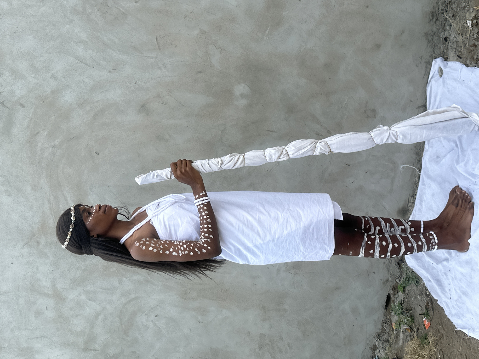 In the city of Badagry, Nigeria, a young Black woman stands facing right in profile against a textured concrete wall. She wears a simple white dress that wraps around her body, and a decorative headband adorns her head, featuring shell-like elements. Her skin is adorned with white paint, applied in dots on her upper right arm and in concentric patterns on her lower legs. White bracelets are visible on her upper right arm, and white paint is applied to her face in a stylized pattern. She holds a long, thick staff wrapped in white fabric and tied with string. The staff rests on a white cloth spread on the ground, which also partially covers the rough, dusty terrain. The lighting suggests an outdoor setting during the day, with a neutral grey background and a hint of dry vegetation in the lower left corner.
