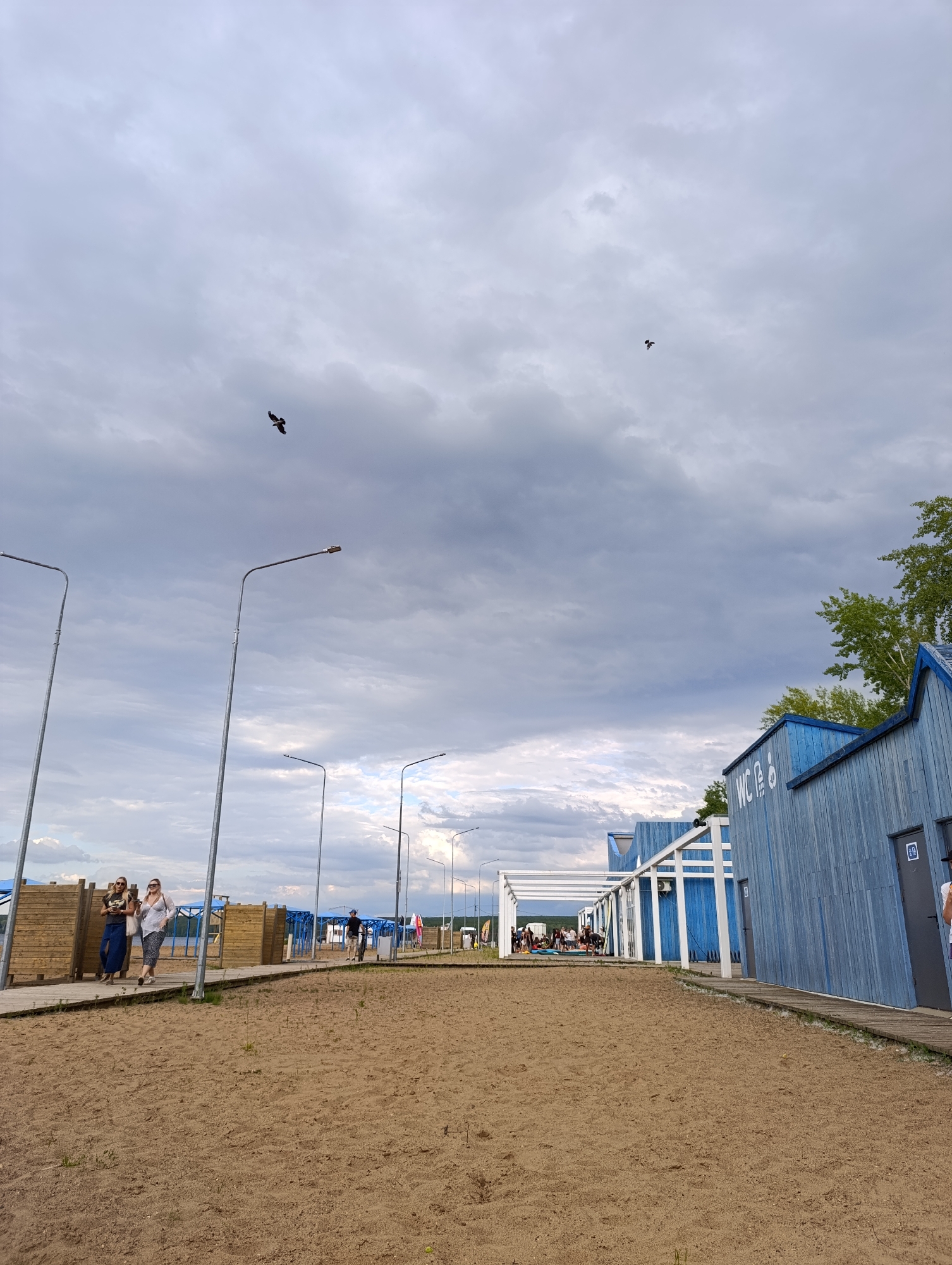 The image depicts a sandy area with a wooden boardwalk and several blue buildings, including structures labeled "WC". The scene is set outdoors under a cloudy sky. In the distance, trees and a body of water are visible, suggesting a recreational area, possibly a beach or park. Several people are present, walking on the boardwalk or gathered in groups. There are also streetlights along the boardwalk. The presence of the "WC" signs and the overall setting implies a public facility in the city of Chaikovsky, Russia. One bird is flying in the sky.
