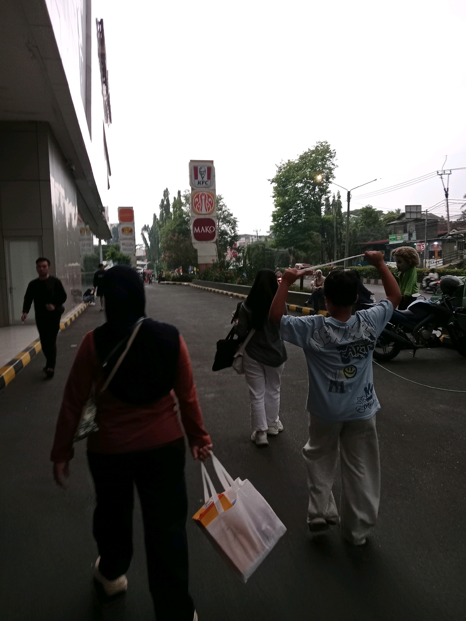 The image depicts a street scene in Cianjur, Indonesia, with a prominent KFC sign visible. Several individuals are walking on the asphalt road. In the foreground, a person wearing a dark hijab and a reddish-brown long-sleeved shirt walks away from the camera, carrying a white shopping bag with orange and yellow accents. To their left, a man in a black long-sleeved shirt and black pants walks towards the camera. Further down the road, another person in a black shirt and pants is visible. In the middle ground, a young person in a blue t-shirt and light-colored pants stands with their arms raised, holding a thin white string. A motorcycle is parked on the right side of the road, and several trees line the background. The sky is overcast, suggesting it might be dawn, dusk, or a cloudy day. The environment appears to be a commercial or urban area, with buildings and signs indicating businesses.