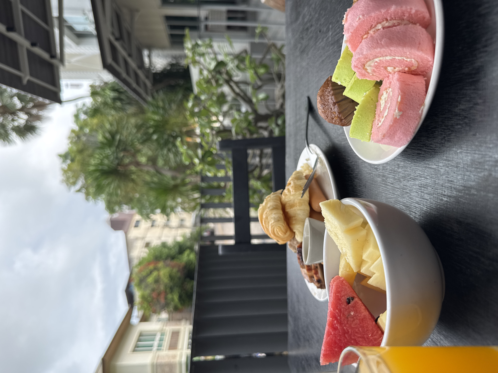 A close-up, eye-level, outdoor shot shows a breakfast spread on a dark, textured table. In the foreground, a white bowl contains slices of red watermelon and yellow pineapple. To the left, a glass of orange juice is visible. To the right, a white plate holds three slices of pink Swiss roll cake, two slices of green cake, and a brown mini muffin. In the background, a white plate holds two croissants and some sausages. Beyond the table, a black metal fence and a black chair are visible. Further in the background, buildings and lush green trees suggest an urban environment, likely in Pattaya City, Thailand, under a cloudy sky.