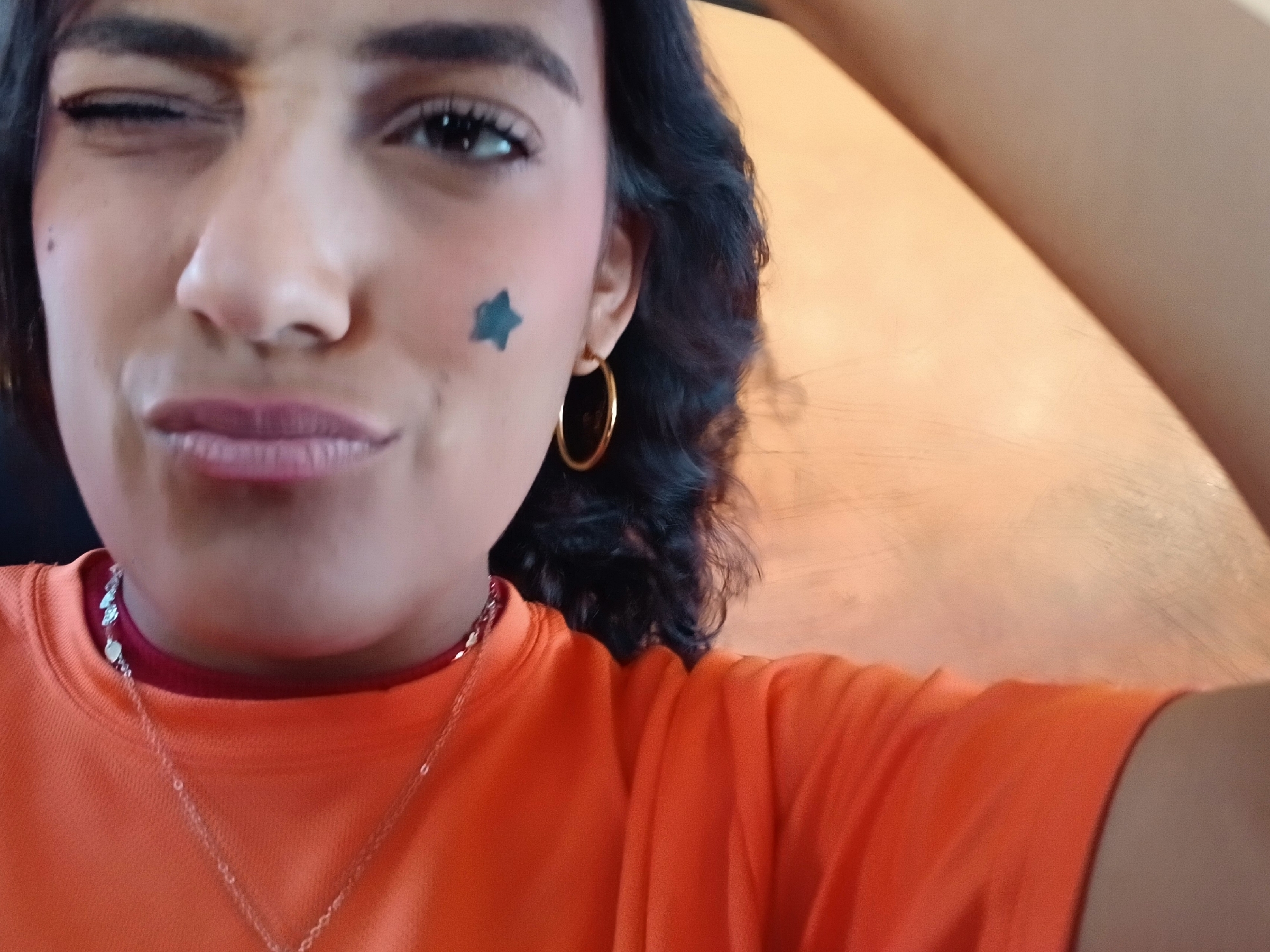 A close-up selfie of a person with dark hair and medium skin tone. The person is winking with their left eye and has a blue star painted on their left cheek. They are wearing a gold hoop earring in their left ear, a silver necklace, and an orange shirt with a maroon collar underneath. The background is blurred, suggesting a shallow depth of field. Based on the lighting and color palette of the background, it seems like the photo was taken indoors. There is no visible information to identify that the photo was taken in the city of Altagracia, Venezuela.
