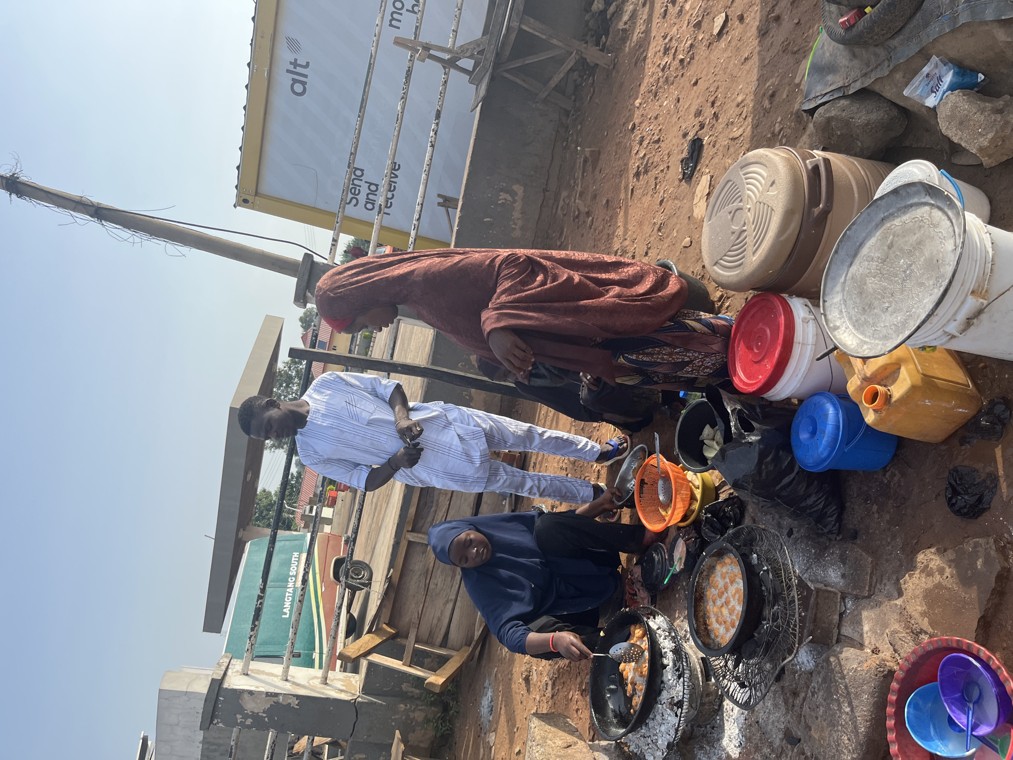 In the city of Gura Top, Nigeria, three individuals are engaged in the preparation and sale of fried food. A woman wearing a blue hijab and dark clothing is crouched down, actively frying food in a large pan placed over burning coals. She uses a slotted spoon to stir the contents of the pan. Beside her, a man in a white striped traditional outfit stands observing the process. To the right, another woman, draped in a burgundy hijab and patterned skirt, is also present, though her specific activity is not as clearly defined. The scene is set outdoors on a dirt ground, with various cooking implements, food items, containers, and a black garbage bag scattered around. In the background, a green and red bus with the inscription "LANGTANG SOUTH" is visible, along with a shipping container labeled "alt." The environment suggests a bustling street-side food vending operation.