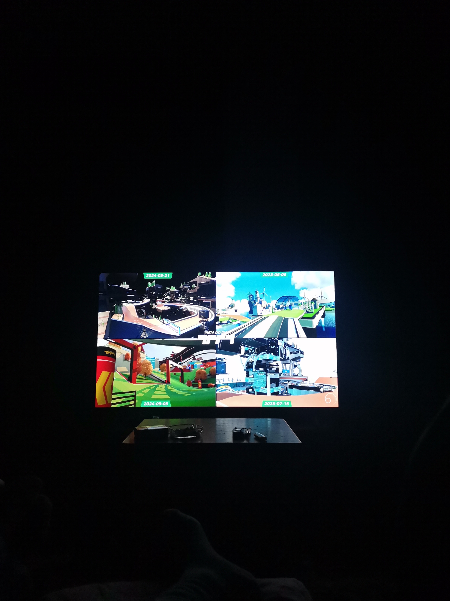 A low-lit indoor scene shows a television screen displaying four different scenes. Each scene has a date overlaid, suggesting a presentation or a game menu. The top-left scene is dated "2024-05-21" and appears to be a futuristic racetrack. The top-right scene is dated "2023-08-06" and shows a bright, futuristic city with windmills. The bottom-left scene, dated "2024-09-05," presents an autumnal landscape with red and orange buildings. The bottom-right scene, dated "2025-07-16," showcases a complex, modern industrial structure. Below the television, a dark surface holds a few small, indistinct objects, possibly remote controls. The remainder of the scene is obscured by darkness, with parts of a person’s legs and feet visible in the foreground. The location is unknown.