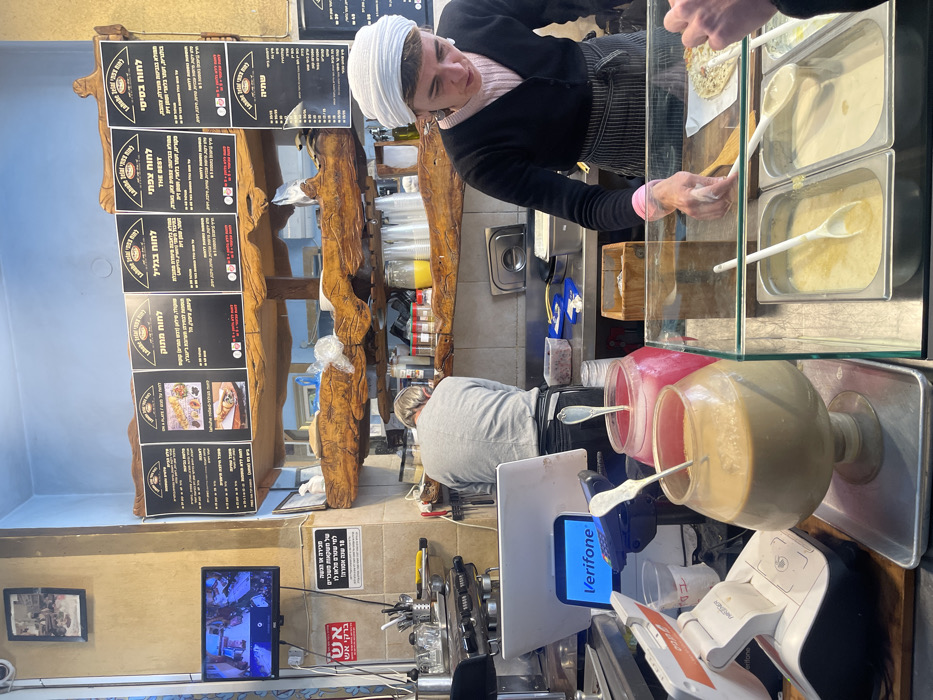 The image captures the interior counter and preparation area of an eatery named "Labane Zefati" (Safed Labaneh) in Safed, Israel.
In the foreground, a white point-of-sale system is positioned on the left, next to a black Verifone payment terminal displaying "Verifone" on its screen. To the right, two large, footed glass dispensers are visible on a metal tray; the left dispenser contains a beige liquid, and the right contains a pink liquid, both with silver serving spoons. Adjacent to these, a glass display case holds two stainless steel inserts filled with pale, possibly creamy, food items, each with a white serving spoon. A piece of flatbread or pita rests on a wooden surface within this display. A customer's hand is visible reaching towards the display from the bottom right.
A female staff member, wearing a white head covering, a black cardigan over a pink undershirt, and a striped apron, stands behind the display, actively reaching into it with her right hand. Behind her, a male staff member in a grey long-sleeve shirt is seen from the back, engaged in tasks at a stainless steel counter. This counter area contains various jars, spices, and kitchen implements, including a plastic container with dark berries or olives.
The background wall features a rustic wooden structure supporting multiple black menu boards with Hebrew text, some indicating "Labane Zefati" and English titles like "ON A PLATE / GOOL" and "THE BEST". Above these menus, the wall is painted light blue; below, it is beige. On the left wall, a small black flat-screen television displays content, with a framed photograph mounted above it. Below the television, a red sign with Hebrew text and a white sign with black Hebrew text are affixed to the wall.