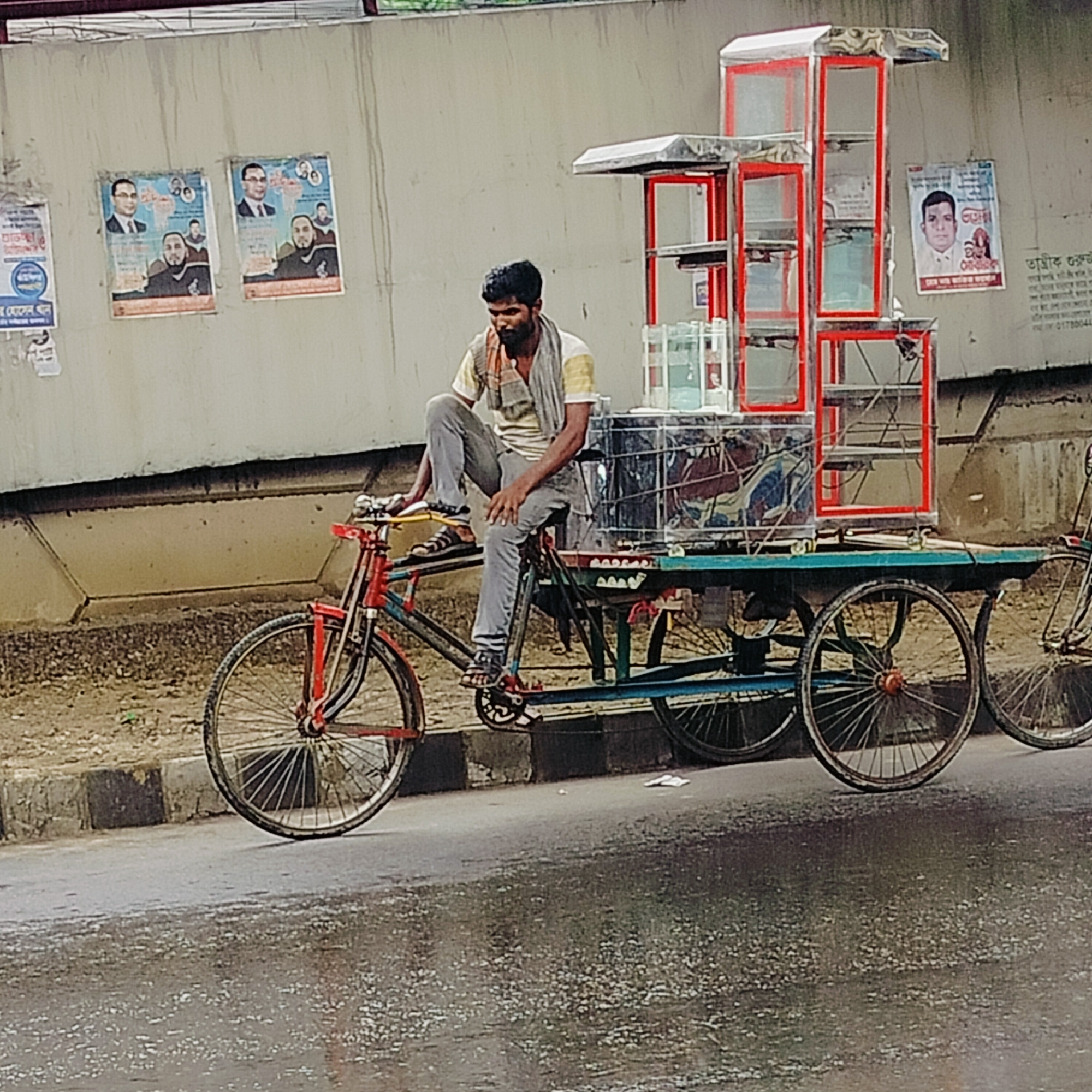 A man is sitting on a tricycle rickshaw on a wet street in Dhaka, Bangladesh. The rickshaw is loaded with several glass containers housed within a red-framed display structure. The man is dark-haired, wearing a light-colored t-shirt, gray pants, and sandals, with a scarf draped around his neck. He is in a pensive pose, with his hand near his face.
The tricycle is multi-colored, with a red front wheel and a blue frame supporting a green platform where the containers sit. The platform has three additional wheels. To the right of the rickshaw, another bicycle is visible, partially out of frame.
Behind the rickshaw, there is a concrete wall covered with several posters. The posters feature images of men, presumably political or religious figures. The street is wet, indicating recent rain.