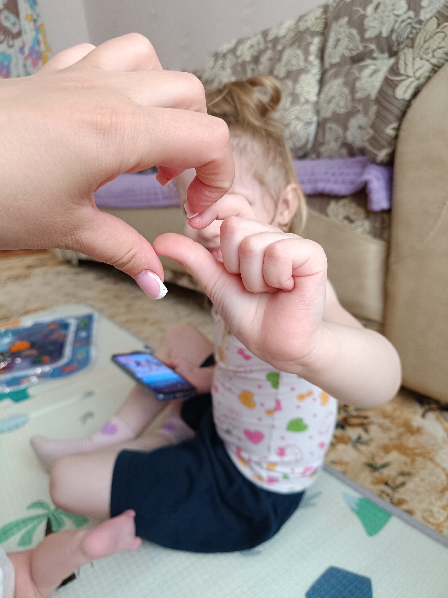 A small child sits on a playmat in a room, facing away from the camera with blonde hair tied up in a small bun. They wear a white top with heart and leaf patterns and navy blue shorts. The child holds a smartphone. An adult hand with a manicured nail forms half of a heart shape in the foreground, and the child's hand makes the other half. A playmat with a pattern of trees and geometric shapes is visible on the floor. A box, possibly a toy box, with marine-themed images lies nearby. The background includes a couch and a patterned blanket or throw. The specific location within Tobolsk, Russia cannot be determined from the image alone.