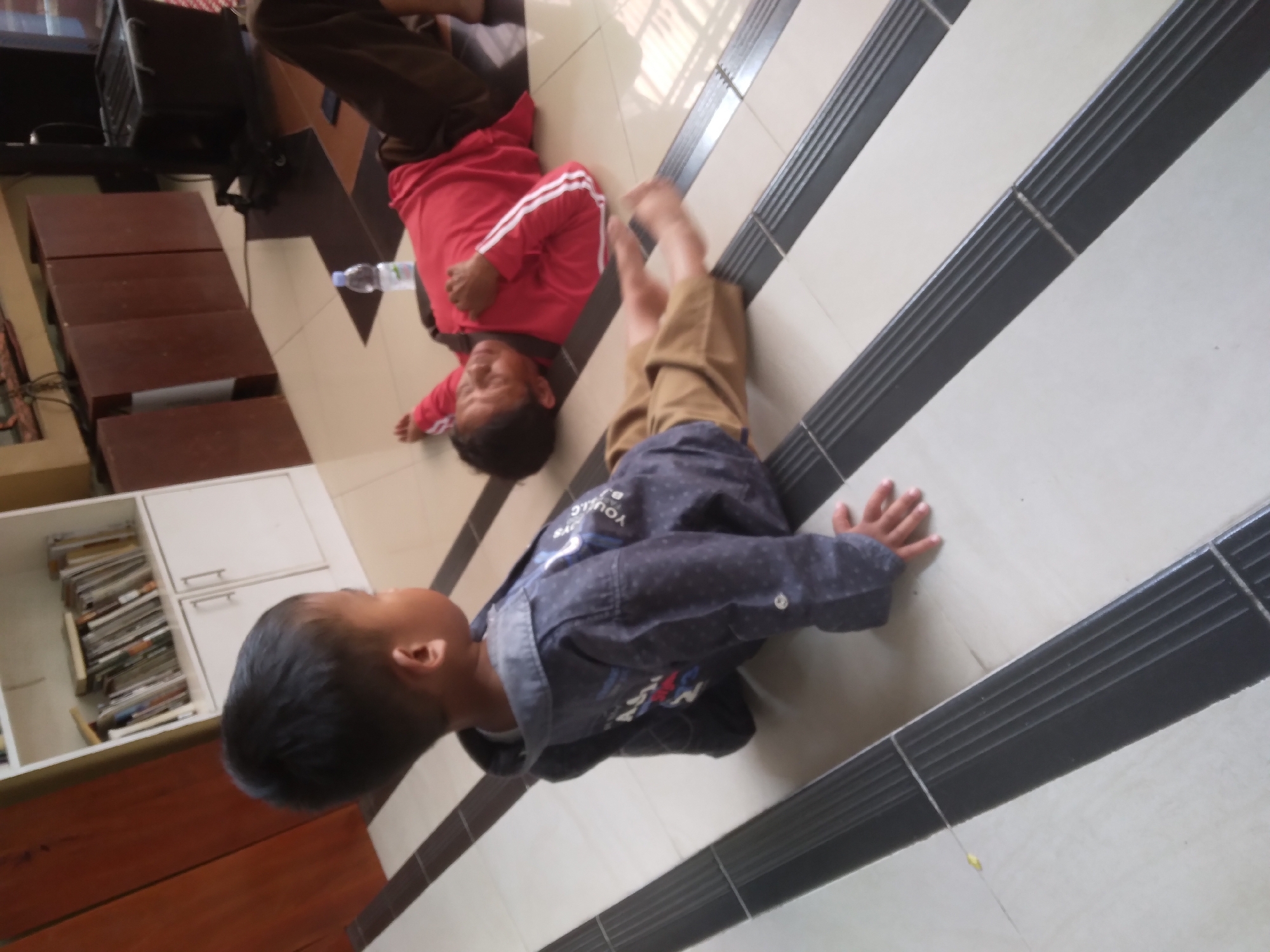 In Desa Pamijen, Kecamatan Sokaraja, Indonesia, two young children are on a tiled floor in what appears to be a room with a bookshelf in the background. One child, on the left, is sitting on their knees and looking to the right. They are wearing a dark-colored collared shirt with a pattern and light-colored pants. The other child, on the right, is lying on their back with their arms outstretched and legs bent. They are wearing a red long-sleeved shirt with white stripes and dark pants. A plastic water bottle is visible near this child. The flooring is a repeating pattern of beige and dark gray tiles with raised black strips. In the background, a white bookshelf filled with books and folders is present, along with some wooden furniture and electronic equipment.