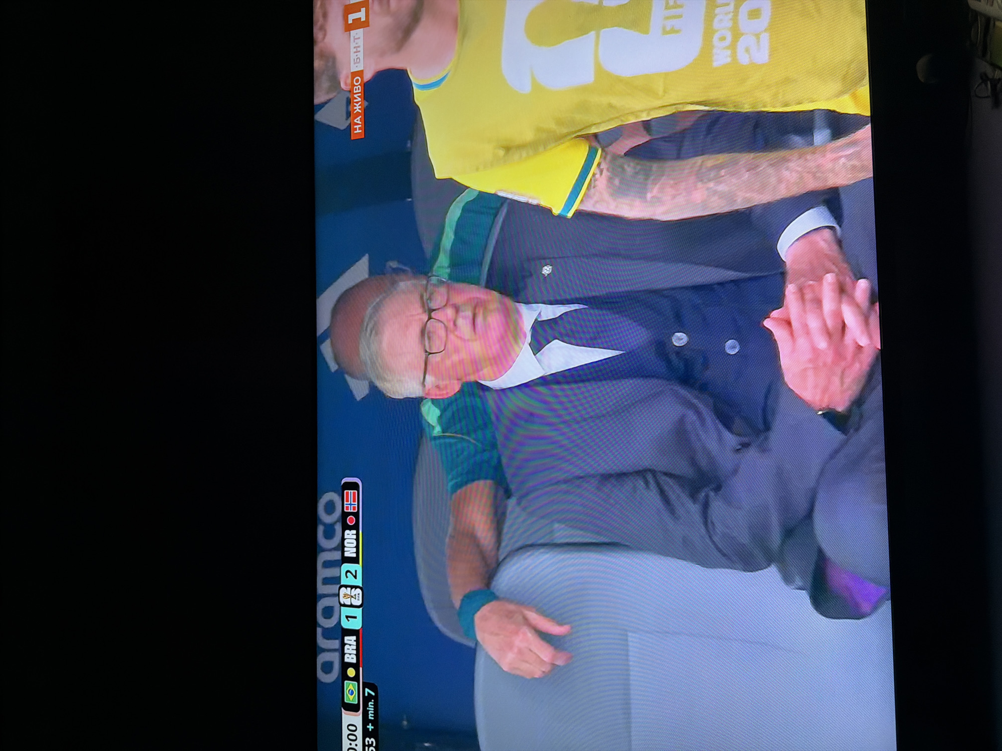The image shows a screenshot from a soccer match broadcast, displayed on a television. In the foreground, a man in a dark suit and tie with glasses is seated, looking slightly to his left. His hands are clasped in his lap. To his right, another person, partially visible and wearing a yellow shirt, is also seated. Behind these individuals, a person in a green shirt with a blue wristband is leaning on a seatback, their arm extended. A scoreboard graphic at the bottom of the screen shows "0:00", flags of Brazil and Norway, and the score "BRA 1 2 NOR". Another graphic at the top right indicates "НА ЖИВО Б.Н.Т. 1". The background consists of blue seating and a dark ceiling. The image is not from the city of Ognianovo, Bulgaria; it is a televised sporting event.