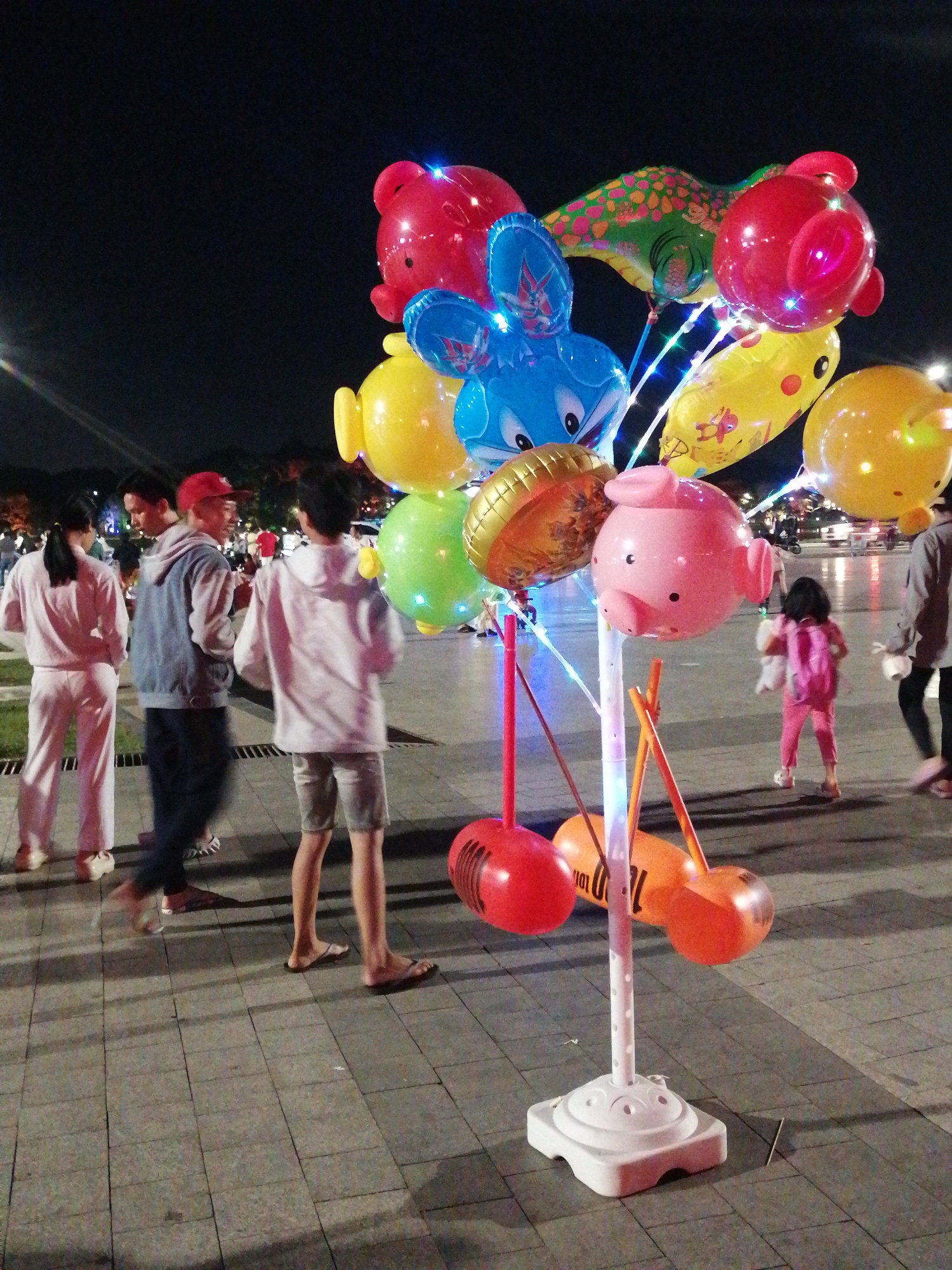 The image shows a street scene at night in Da Lat, Vietnam, likely a pedestrian walkway or plaza. In the foreground, a stand holding a cluster of brightly colored balloons is prominently displayed. The balloons are shaped like various cartoon characters and animals, including pigs, bears, a bunny, and a fish-like creature. Some of the balloons are illuminated with strings of small, colorful lights.
Several people are visible in the image, walking and interacting. On the left, three individuals are walking away from the camera. The person closest to the camera on the left is wearing a pink tracksuit. Next to them is a male wearing a denim jacket and a red baseball cap. The third person in this group is a male wearing a white hoodie and shorts. Further to the right, a young girl in pink is walking away from the balloon stand, carrying a white object. Another adult figure is visible in the far right, walking away from the camera, wearing dark pants and a grey top. The ground is paved with square tiles. In the background, the scene suggests an urban setting with distant lights and some blurred figures, indicating other people or activities. The overall atmosphere is festive, likely a night market or a public gathering area.