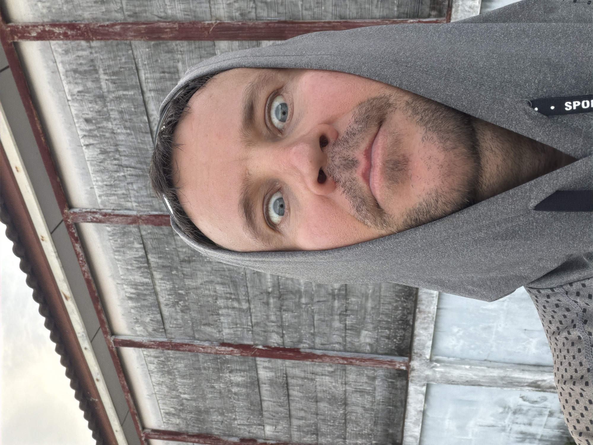 A medium-close-up selfie shows a man wearing a grey hooded sweatshirt. The man's face is centered in the frame, with his eyes wide open and looking directly at the camera. He has short brown hair, a mustache, and a light beard. Behind him is a weathered wooden building with red vertical beams and a corrugated metal roof. The building appears to be located in Baan Mae Nam, Thailand. The overall impression is that of a candid, informal snapshot taken outdoors.