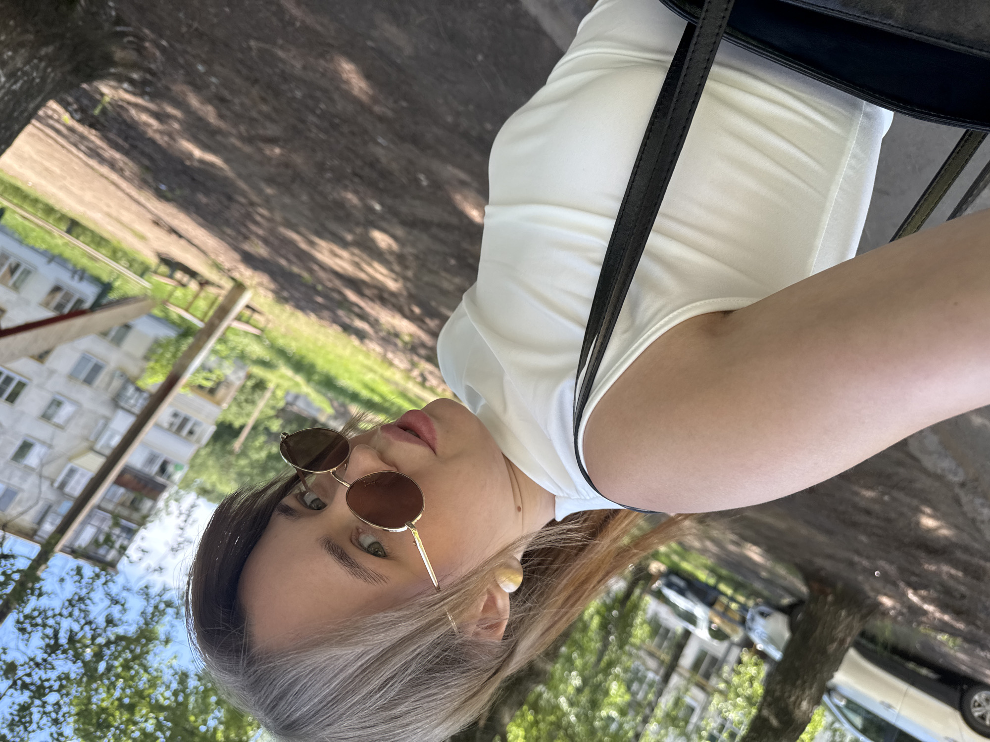 A woman wearing sunglasses and a white shirt is standing outside in Chaikovsky, Russia. The background includes trees, grass, buildings, and cars. The woman is positioned to the right of the frame, her head angled slightly. A black bag strap is visible over her shoulder.