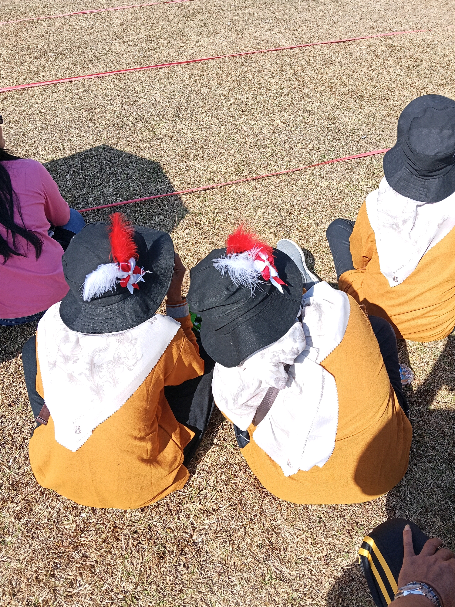 The image depicts an outdoor scene on a dry, grassy field, possibly at a park or an open area in Bendosewu, Indonesia. Three individuals are visible, all seated on the ground, facing away from the camera. The two individuals in the foreground are wearing similar outfits: black bucket hats adorned with red and white feathers, and orange tops with white patterned fabric draped over their shoulders. Their lower bodies are clad in dark pants. To the right, a third individual is also seated, wearing a black bucket hat and an orange top with a white draped fabric. In the background, a person wearing a pink long-sleeved shirt and blue jeans is also seated. Red tape or ribbon is laid out on the ground in parallel lines, suggesting some sort of demarcation or event setup. The ground is covered in dry, light-brown grass, and shadows cast by the individuals and an unseen object are visible. The overall impression is of a gathering or event taking place under sunny conditions.