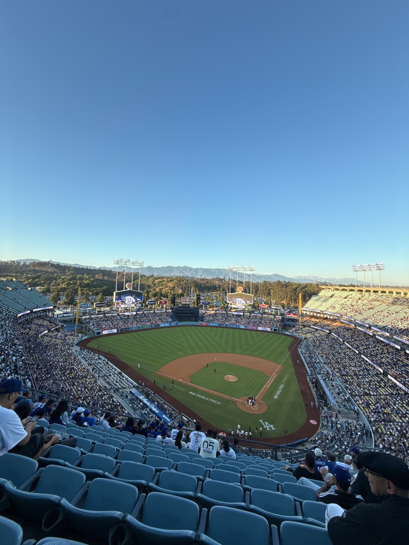 The image depicts a baseball game at Dodger Stadium in Los Angeles, California. The stadium is filled with spectators in the seats, and a game is in progress on the field. Mountains are visible in the background. The sky is clear and blue.