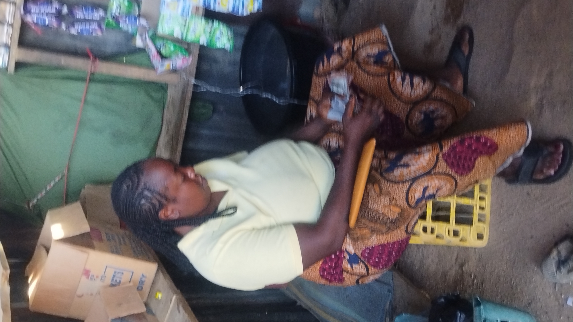This media file captures a seated woman engaged in a transaction or counting money within what appears to be a small, informal shop or market stall. The image is rotated 90 degrees counter-clockwise.
The primary subject is a Black woman with braided hair, wearing a light yellow short-sleeved top and a patterned wrapper or skirt in shades of brown, red, and blue/black. She is seated on the ground, with her legs extended and resting on a yellow plastic crate. She wears dark sandals. In her hands, she holds several light-colored objects, which appear to be banknotes or small pieces of paper, seemingly counting or organizing them. A long, yellow, cylindrical object rests on her lap.
The setting suggests a humble retail environment. To the viewer's left (behind the woman if oriented correctly), there are cardboard boxes stacked, one partially visible with the text "KET3 DRY" and another with "BOS". Behind her, a makeshift shelf system constructed from wooden planks holds various packaged goods in colorful wrappers. A green fabric or tarp serves as a backdrop. To her right, a black basin or bucket is visible on the ground. The ground itself is uneven, appearing to be dirt or rough concrete. The lighting is indirect, suggesting daytime. The woman's expression is neutral and focused on her activity.