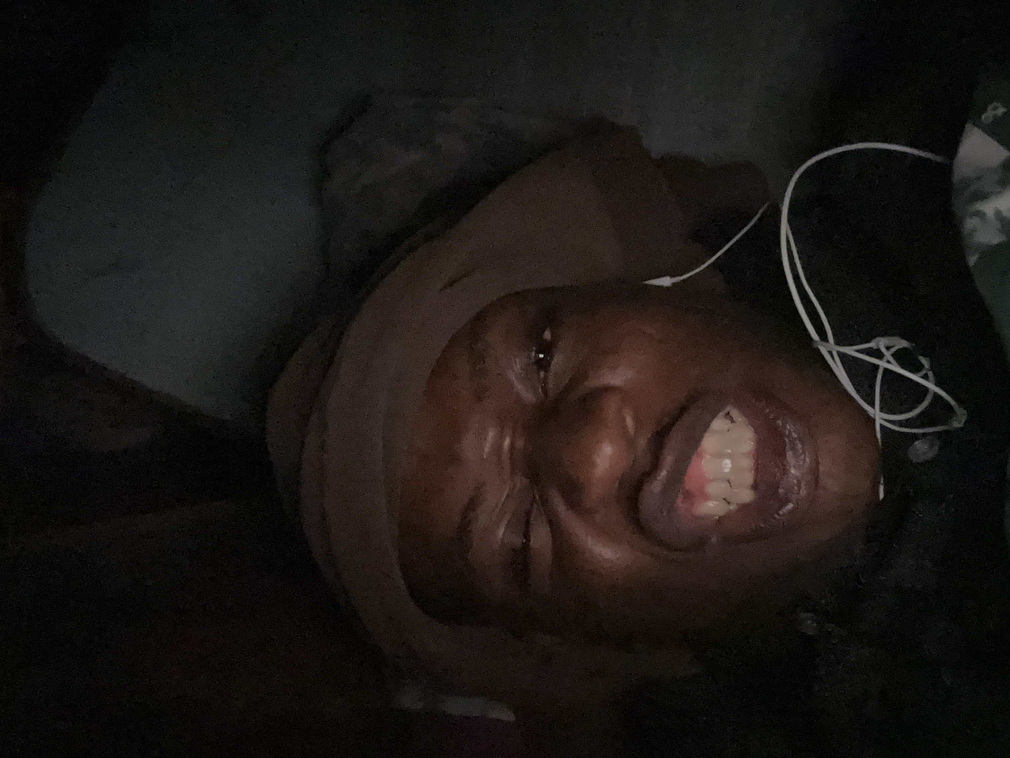 A person is lying down in a dimly lit setting, possibly indoors given the presence of a pillow or cushion under their head. The person is wearing wired earphones. The specific city and context are unknown.