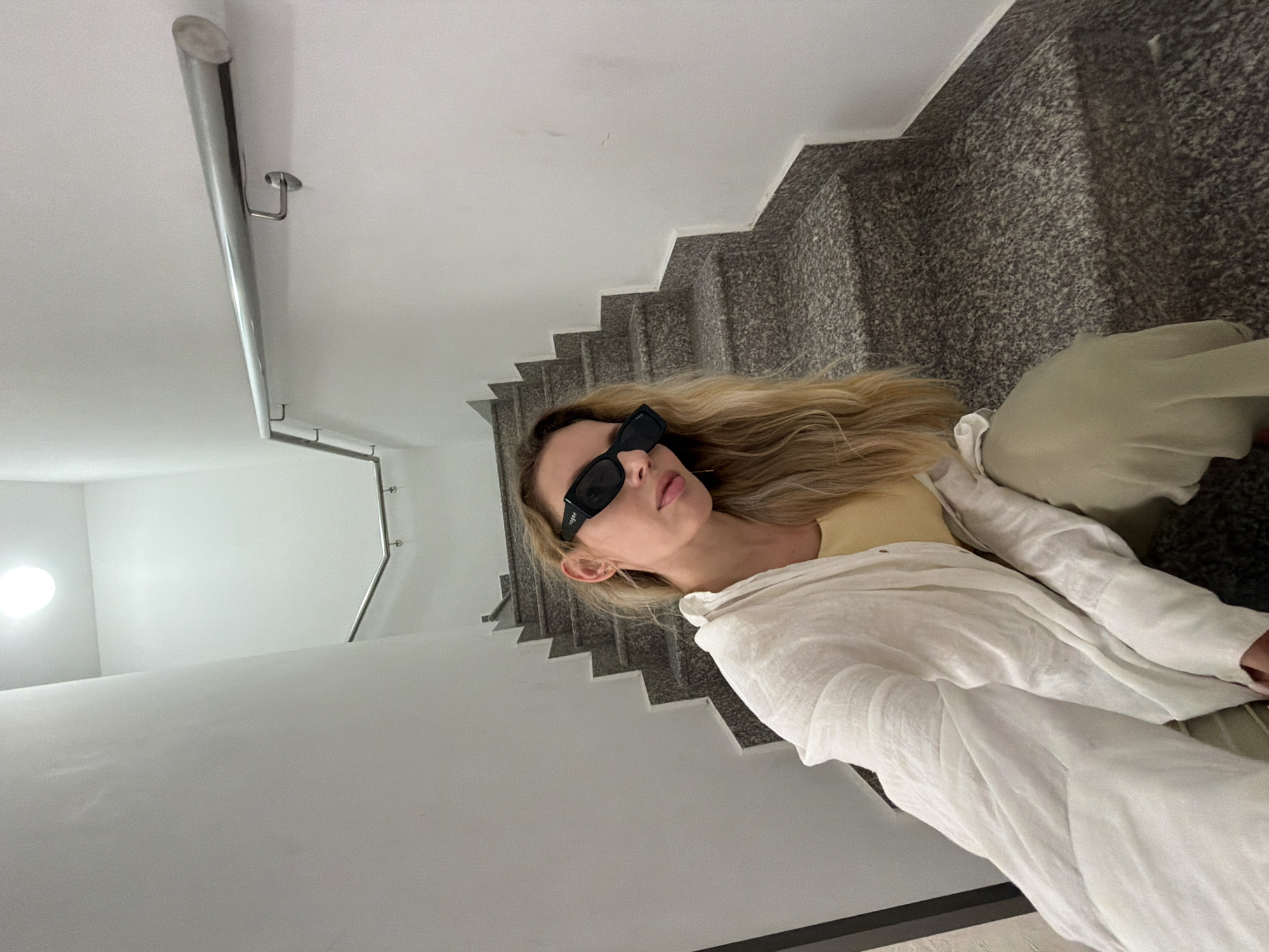 A woman with blonde hair and sunglasses is sitting on a granite staircase in a stairwell. She is wearing a yellow top and a white open shirt. The stairwell has white walls and a metal handrail. The image appears to be a selfie taken in Budva, Montenegro.