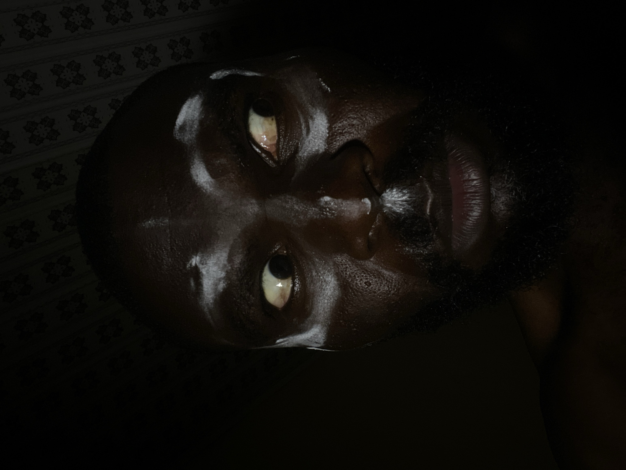 The image is a close-up, low-angle shot of a dark-skinned man's face with a beard. The man is lying on his side, with his head resting on a surface with a patterned background. The man's eyes are open, and there is a sheen of moisture or oil on his face. The image is dark, with only the man's face and the background visible. The scene appears to be indoors. There is no information about the location in the city of Gura Top, Nigeria.