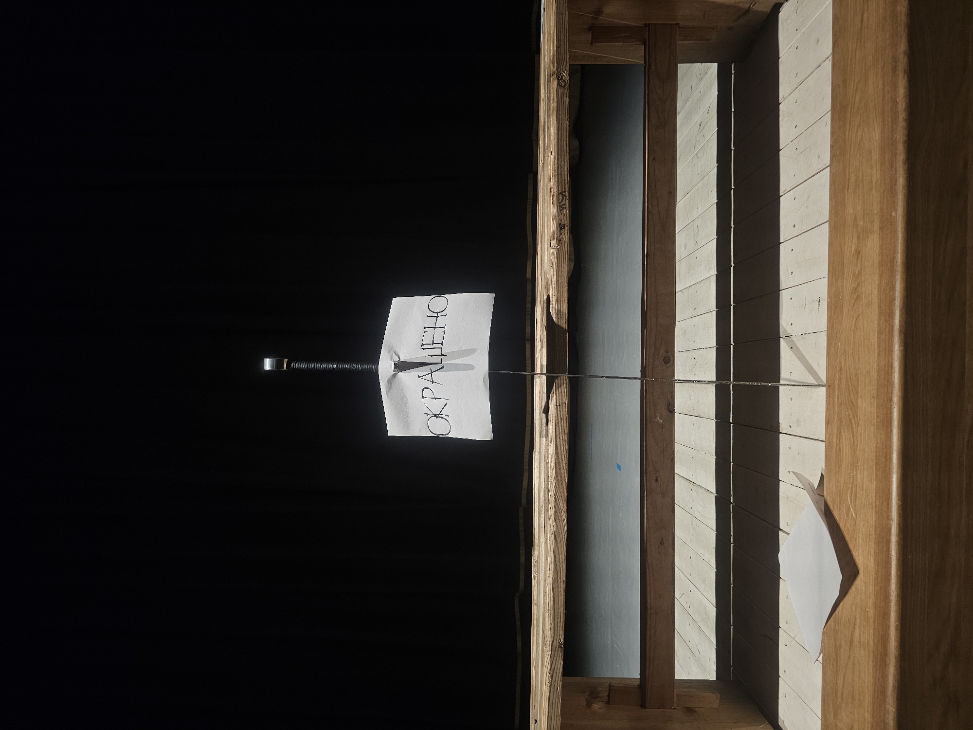 The image depicts a minimalist stage setup with a dark backdrop. A piece of paper with black handwritten Cyrillic text, "ОКРАШЕНО," meaning "painted" or "dyed," is affixed to a metal pole with a nail. The pole extends vertically from a wooden structure that resembles a bench or a platform, supported by wooden beams. The foreground features a wooden surface with a second piece of paper lying on it. The lighting creates strong shadows, suggesting a theatrical or artistic presentation. The scene does not provide any specific geographical indicators that would place it in Tosno, Russia, or any other particular city.