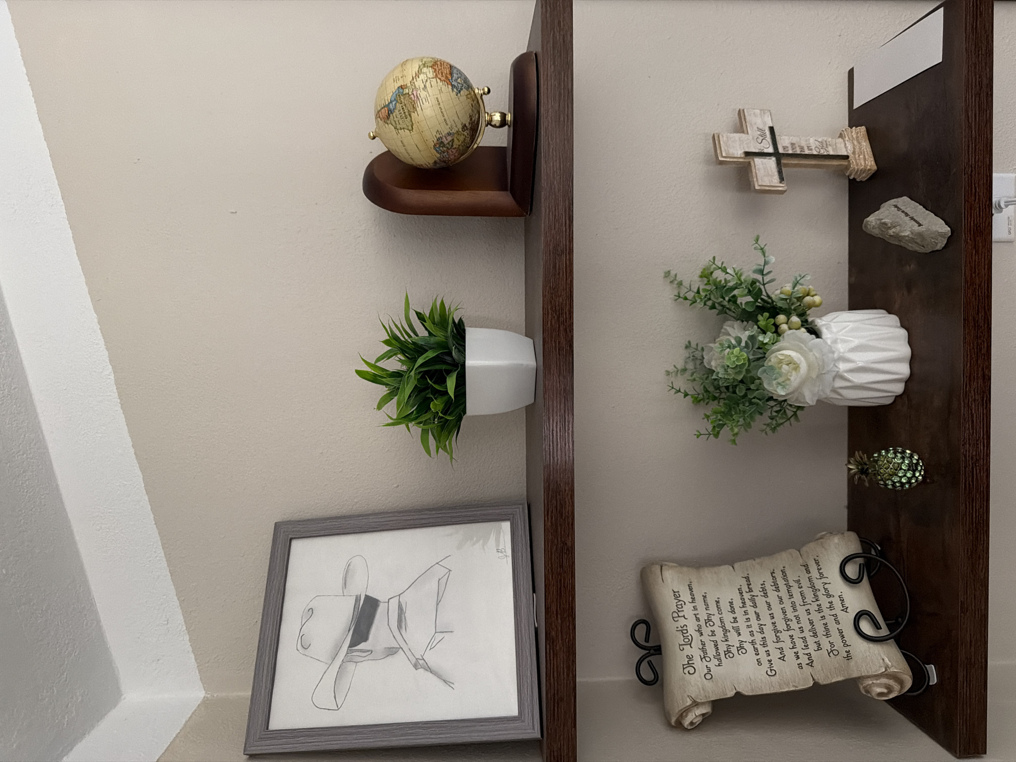Two dark wood shelves are mounted on a light beige wall. The top shelf holds a framed pencil drawing of a cowboy hat and shoulders, a white pot with a green artificial plant, and a small globe on a wooden stand. The bottom shelf displays a scrolled parchment with "The Lord's Prayer," a small, jeweled pineapple figurine, a white vase with an artificial floral arrangement, a small rough grey stone, and a wooden cross with the word "Still" at the top and text below. A white electrical outlet is visible on the wall below the bottom shelf. The scene is located in Wellford, United States of America.