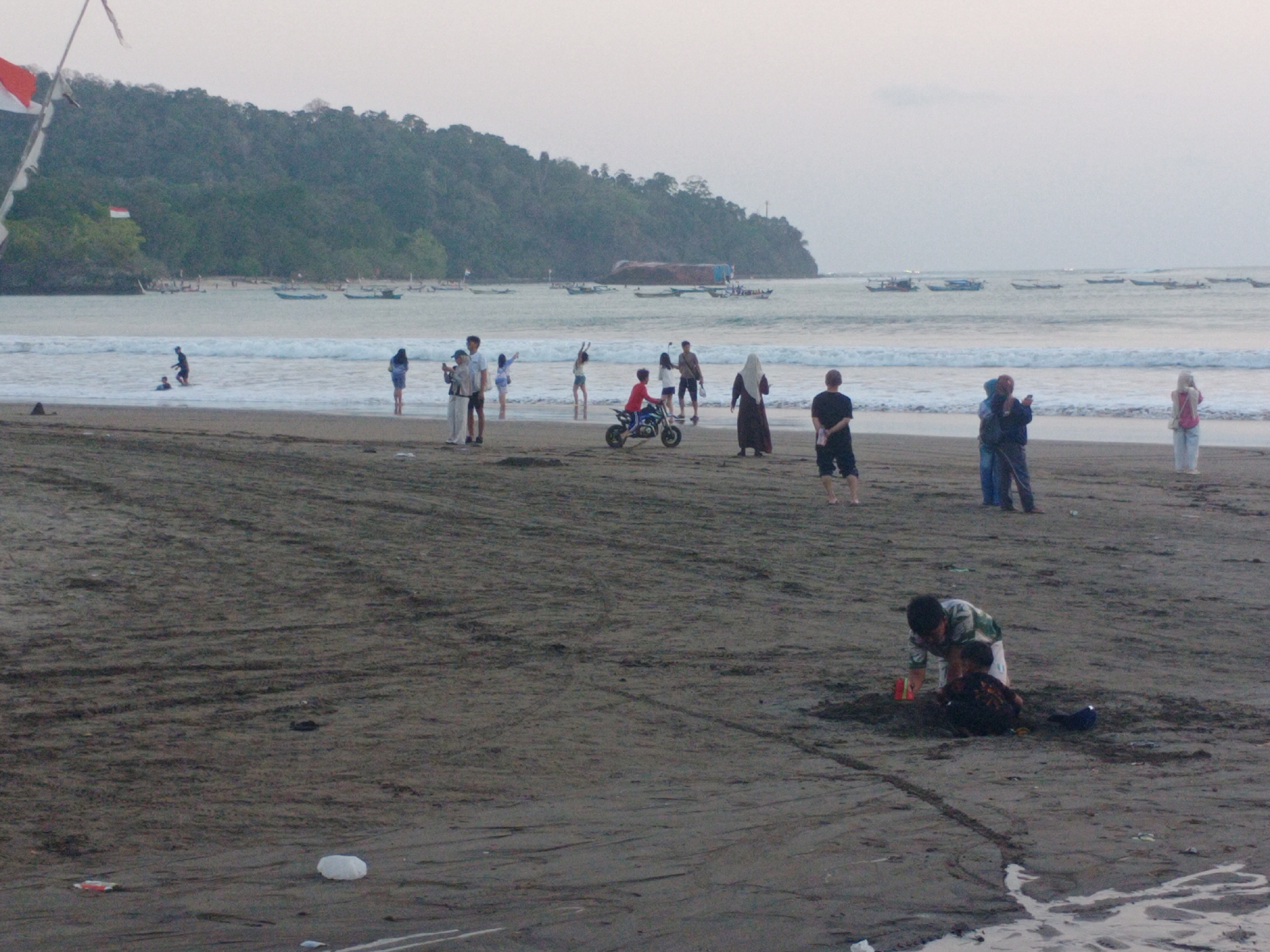 The image depicts a beach scene at Desa Pagandaran, Indonesia. Several people are present on the sandy shore and in the shallow water. In the background, lush green hills rise behind the beach. The sea is calm with gentle waves, and several fishing boats are visible in the distance.
On the beach, a group of individuals are scattered. Some are standing and observing the surroundings, while others are engaged in activities. Closer to the foreground, two people are interacting with the sand, one appearing to be digging or playing. Further down the beach, a person is seen riding a small motorbike.
The environment is a typical beach setting with sand, water, and vegetation in the background. The sky is overcast, suggesting it might be early morning or late afternoon. The overall impression is one of leisure and casual activity at a coastal location.