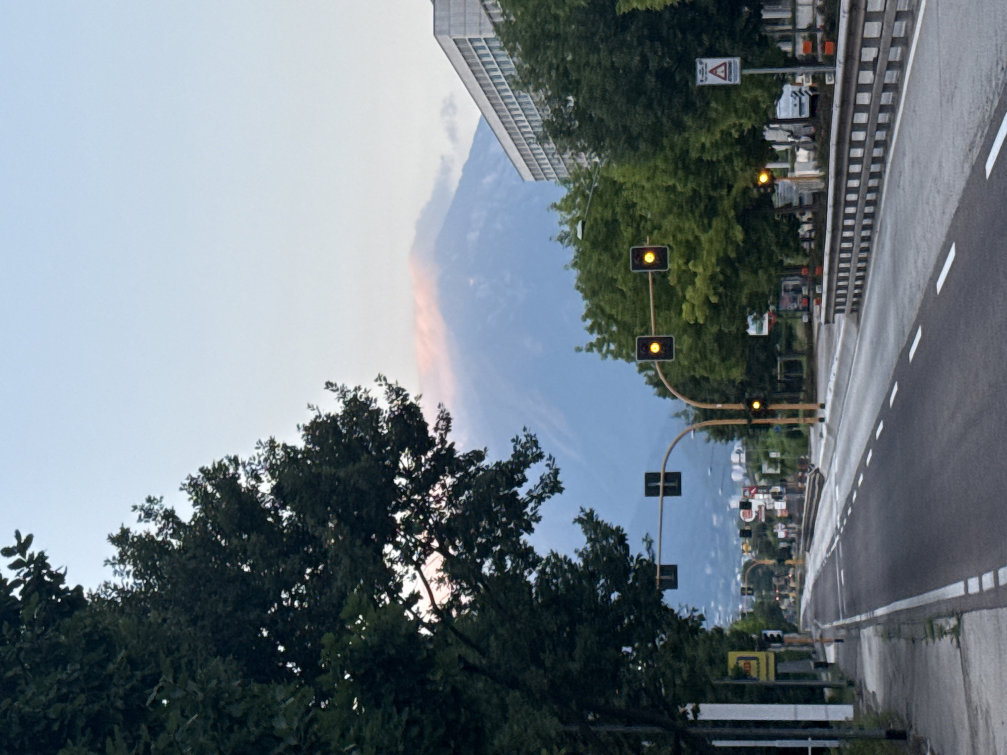 The image depicts a street scene in Trento, Italy, at dusk or dawn. A paved road stretches into the distance, lined by trees on the left and a guardrail on the right. Traffic lights with yellow illumination are visible at intersections. A large, modern building is situated on the right side of the road. In the background, a mountain range is partially obscured by clouds. The sky is a soft gradient of blue and pink. The street appears to be relatively empty, with no people or vehicles prominently featured.