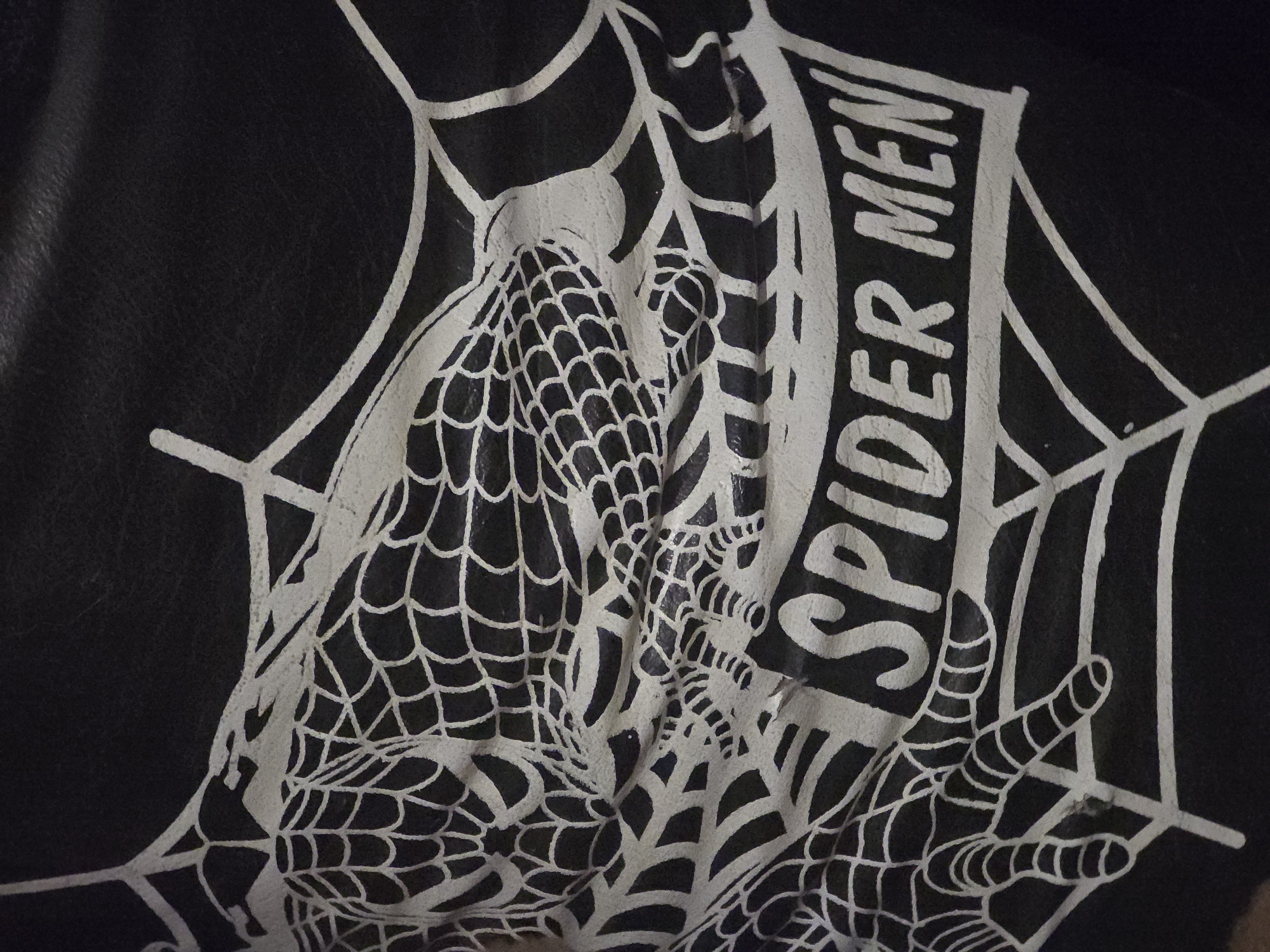 A close-up, high-angle shot of a black surface with a white graphic design is presented. The design depicts a stylized spiderweb with a spider in the upper left quadrant, rendered in a somewhat abstract manner with intersecting lines. Below the spider and web, a banner-like shape contains the words "SPIDER MEN" in bold, white, sans-serif lettering. The surface appears to be textured and slightly creased, suggesting it might be fabric or a similar flexible material. The lighting casts soft shadows that highlight the raised lines of the graphic. No city or environmental context is visible.