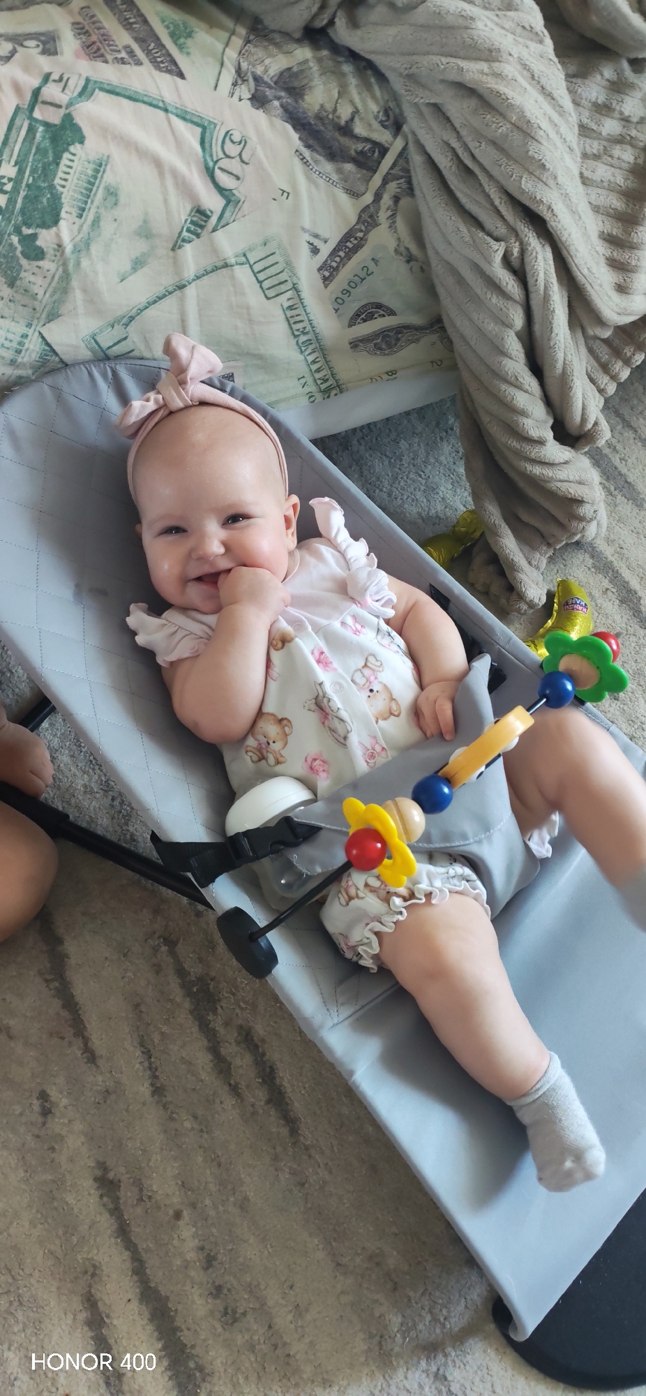 A baby girl is lying in a gray baby bouncer seat. The baby is wearing a pink headband with a bow and a light-colored outfit with teddy bear patterns and ruffles. She is smiling and has her right hand near her mouth. A colorful toy hangs across the front of the bouncer. The bouncer is on a patterned carpet. In the background, there is a bed with a patterned blanket and a beige, textured blanket. The location and city of Noviy, Russia, cannot be determined from the image.