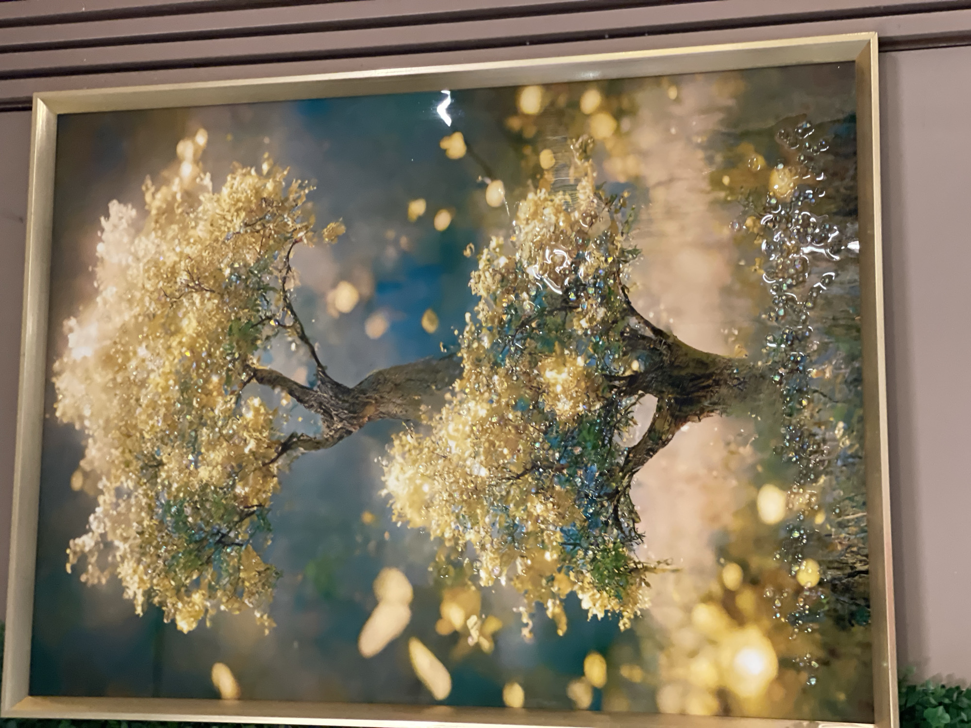 A framed picture of two stylized trees is presented. The trees are depicted in a hazy, bokeh-filled scene, with golden light effects suggestive of sunlight filtering through leaves. The tree on the left has primarily golden leaves, while the tree on the right has both golden and green leaves. The foreground and background contain blurred golden spots that enhance the atmospheric effect. The picture is positioned against a wall, with a glimpse of green foliage visible at the bottom right corner. The location in Akwanga, Nigeria, cannot be determined from the visual information in the image.