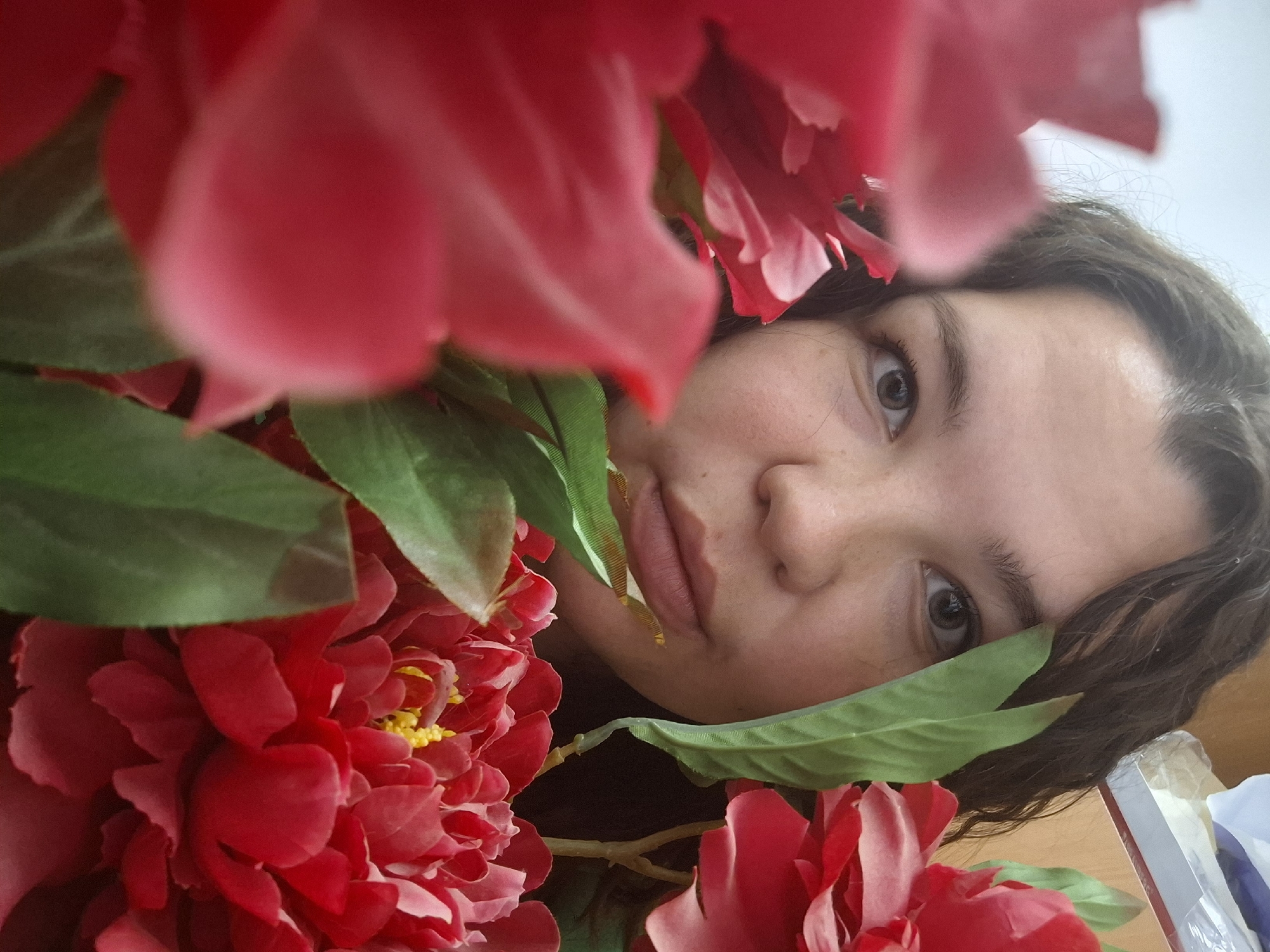 A young woman's face is partially obscured by red flowers and green leaves. The woman has brown eyes and dark, curly hair. The flowers are large and appear to be red peonies or similar blossoms. The leaves are green and interspersed with the flowers. The background is out of focus but appears to be a bright, neutral color. The image is taken from an unusual angle, possibly from above. The city of Unknown is not identified.