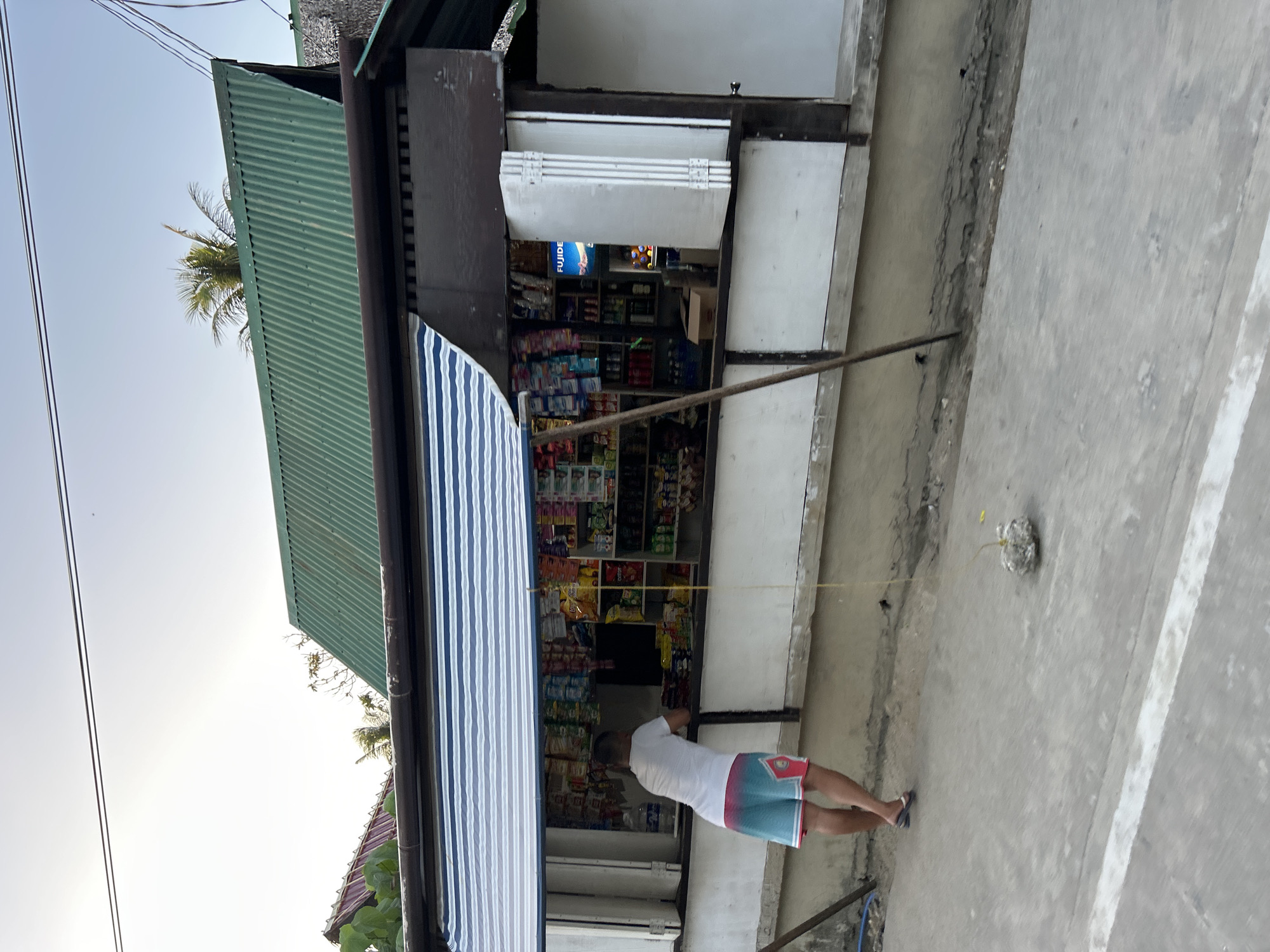This media file captures an outdoor scene in General Luna, Philippines, depicting a local convenience store, commonly known as a sari-sari store, under a bright sky. The image is oriented vertically, showing the store from an angle that includes the street or pathway in the foreground and the sky above.
**Subjects**: The primary subject is a person, seen from behind, standing near the entrance of the store. They are wearing a white t-shirt and blue and pink patterned shorts, with flip-flops on their feet. The store itself is packed with various merchandise, including packaged snacks, drinks, and other goods displayed on shelves. It features a green corrugated metal roof and a blue and white striped awning extended over the front. Power lines are visible in the upper left, and palm trees are seen partially behind the store structure. A long, thin pole, possibly made of metal or bamboo, extends from the store front towards the right side of the frame, with a yellow string attached to it, leading to a small, lumpy, grey object on the concrete ground. The ground is a light grey concrete surface.
**Activity or event**: The person appears to be either attending to the store or making a purchase. The pole and the string leading to the object on the ground might suggest a game or a makeshift setup for an activity, possibly a traditional game or a simple toy.
**Notable details**: The scene takes place during daytime, indicated by the clear, bright sky and soft light, likely in the late afternoon. The architecture of the store, including the corrugated roof and the awning, along with the visible palm trees, are typical of a tropical setting in the Philippines.
**Visible text**: Inside the store, a blue sign with white text "FUJIDE" is visible, likely indicating a brand or product being sold.