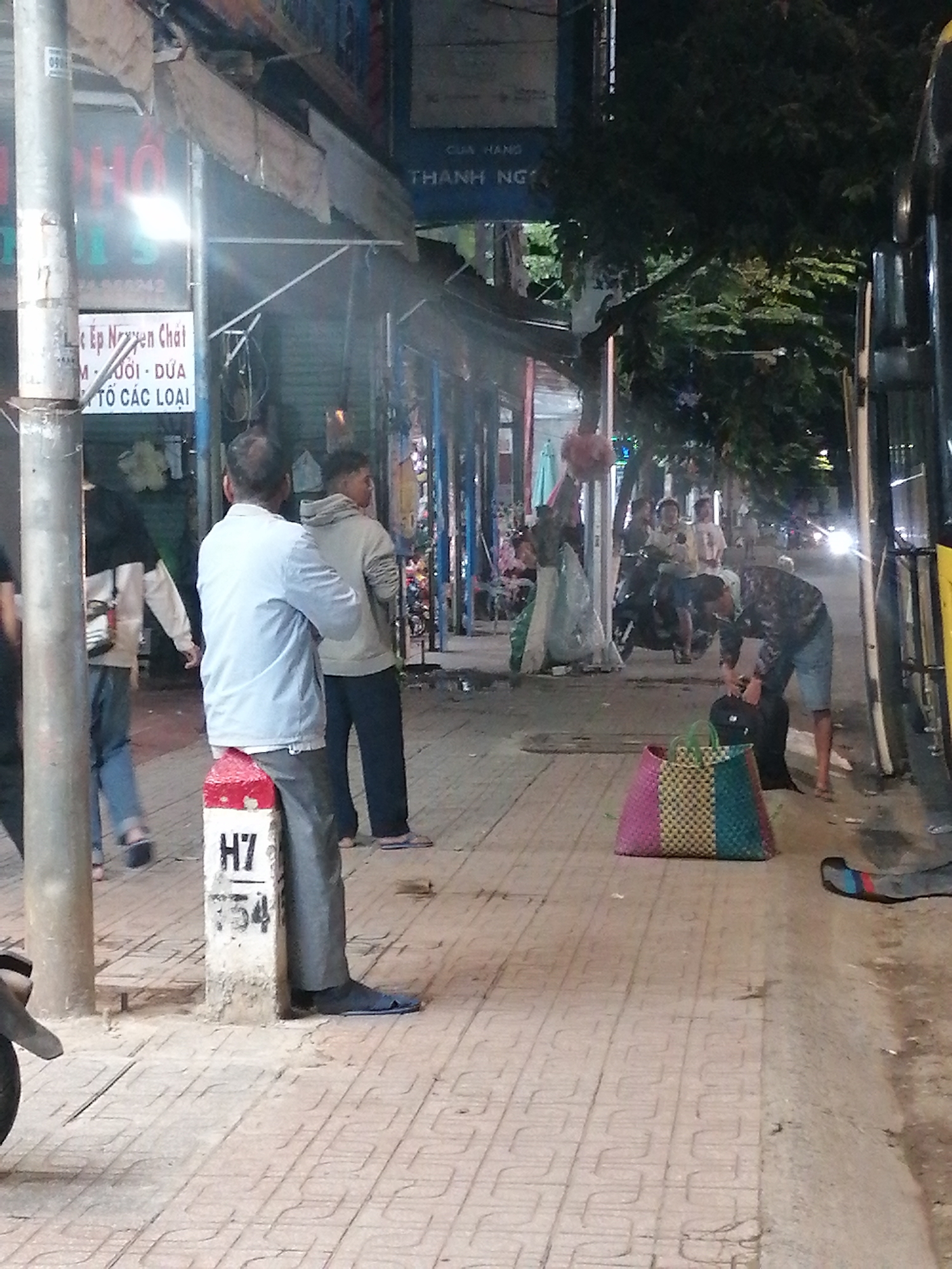 The image depicts a street scene at night in what appears to be Đinh Văn, Vietnam. The foreground shows a paved sidewalk with a distinctive patterned surface. A red and white striped marker post, labeled "H7" and with "754" below it, stands on the sidewalk. To the left of the marker post is a large, colorful woven bag with sections of pink, yellow, and teal. Further down the sidewalk, a person in a dark, patterned shirt and blue shorts is bending over to pick up a black backpack. To the right, the side of a large vehicle, possibly a bus, is visible, with its headlights on.
In the middle ground, several people are on the sidewalk. Two men, one in a light blue shirt and grey pants, and another in a light grey hooded sweatshirt and dark pants, are standing. Behind them, other individuals are visible, some appearing to be on or near motorcycles. A street vendor's stall is set up with awnings and various goods displayed. Signs in Vietnamese are present on the buildings lining the street, one of which reads "THÀNH NG" and another with "LỆ EP NGUYỄN CHAI". The environment appears to be an urban setting with shops and trees. The lighting suggests it is after sunset.