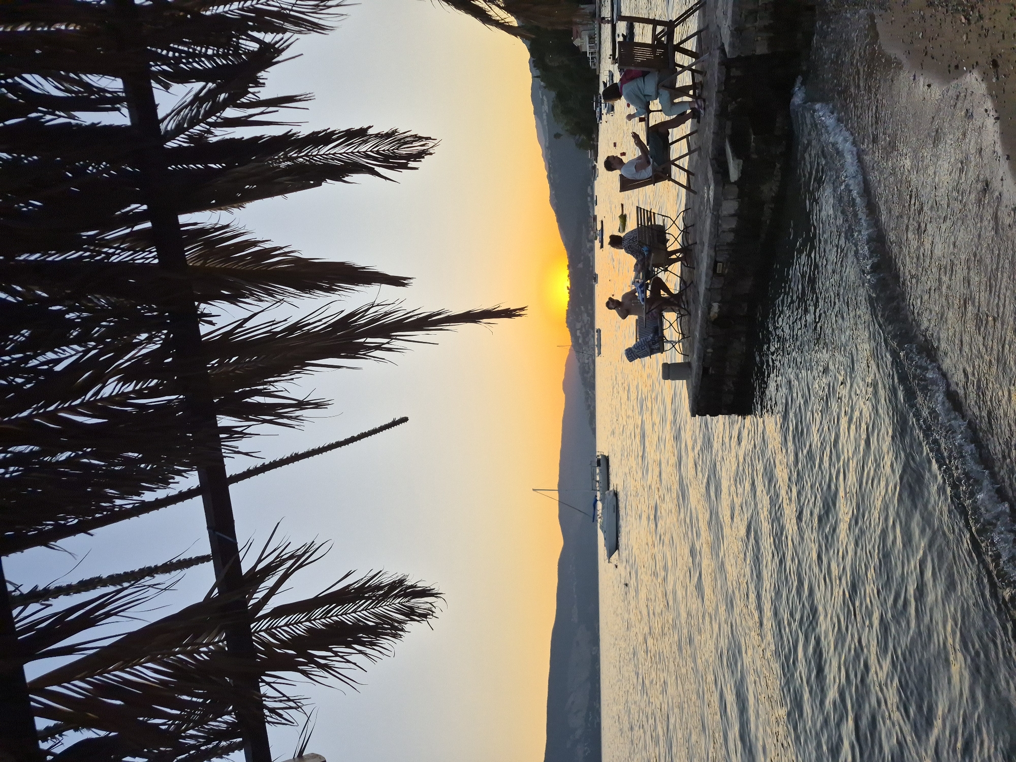 In Kumbor, Montenegro, at sunset, four people are seated at tables on a stone pier extending into calm water. Two individuals are seated at a table on the left, while two others are seated at a table on the right. They appear to be engaged in conversation or dining. The pier is furnished with chairs and tables. Several boats are anchored in the bay, and sailboats are visible in the distance. The sun is low on the horizon, casting a warm orange glow across the sky and water. Palm fronds from an overhead structure hang down, framing the scene. The shoreline is visible on the right, with sand and small pebbles. The overall atmosphere is serene and picturesque.