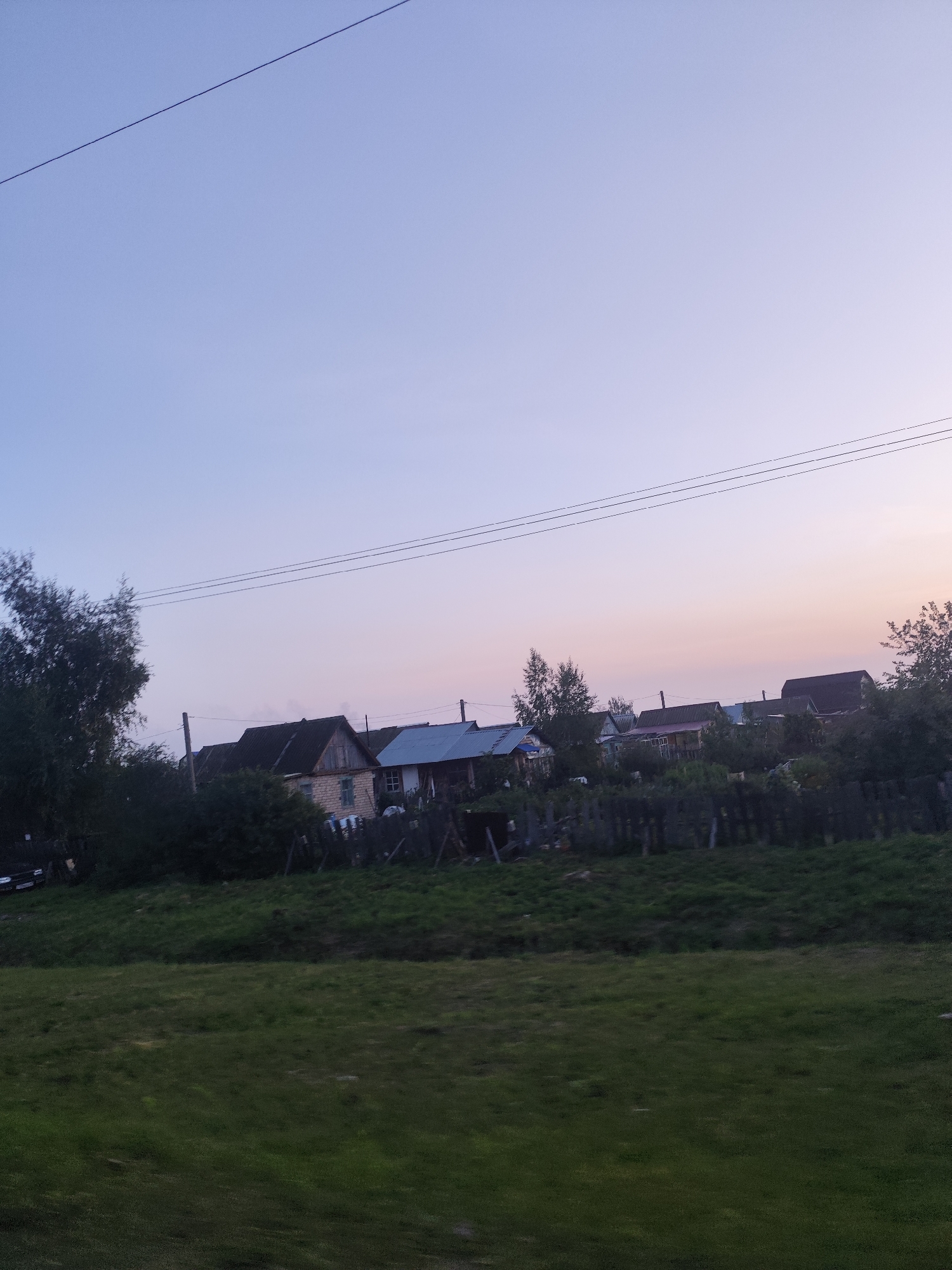 The image depicts a rural landscape under a pale pink and blue twilight sky. In the middle ground, a row of modest houses with dark roofs and light-colored walls are visible, interspersed with trees and a wooden fence. A power line stretches across the sky. The foreground consists of a grassy field. The scene suggests a quiet, undeveloped area. It is not possible to determine the specific location within the city of Petropavl, Kazakhstan, from the provided image.
