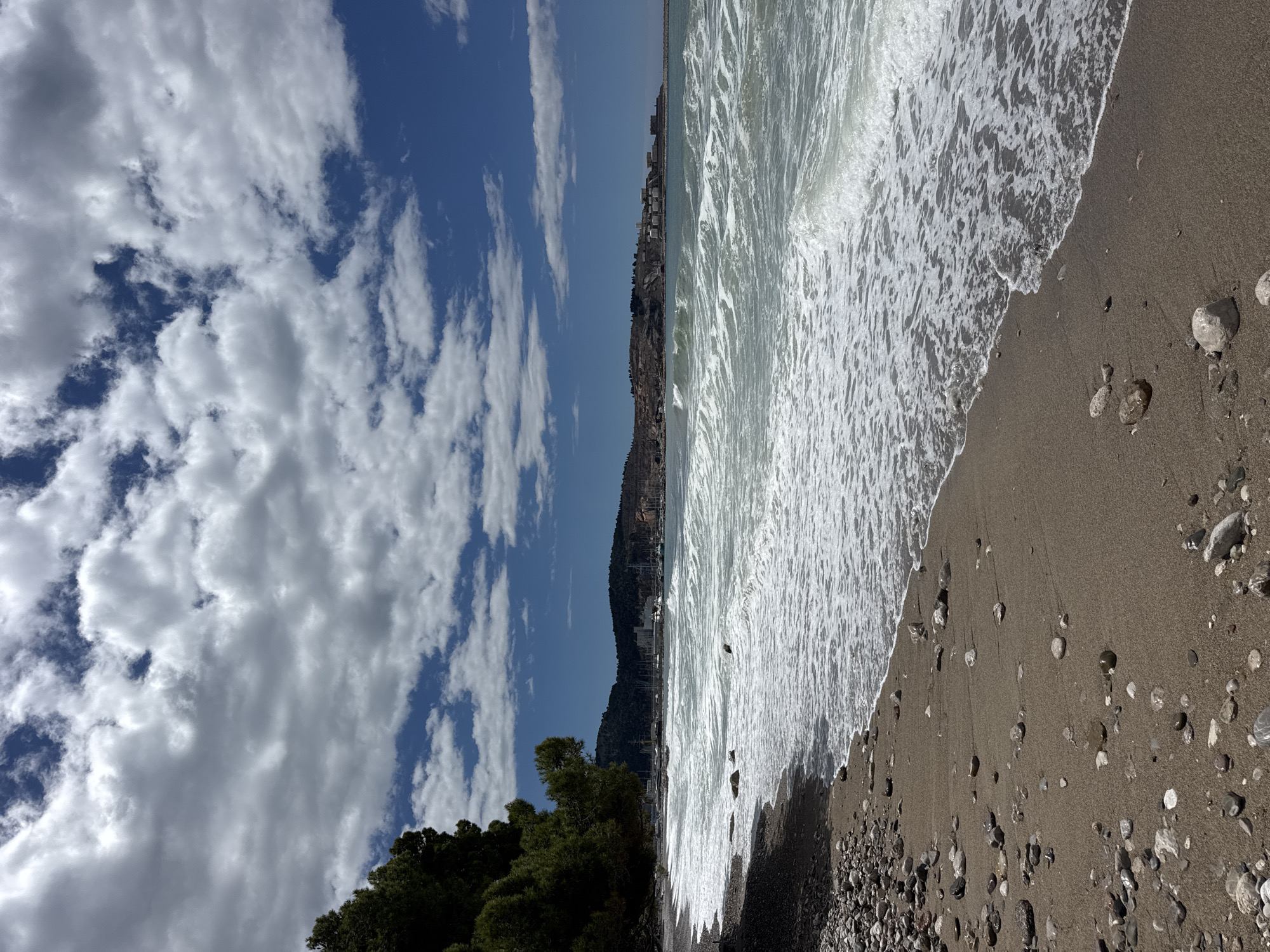 A beach on the Adriatic coast of Bar, Montenegro. Waves wash onto a dark sand and pebble beach. Trees line the left edge of the image, with buildings visible in the distance on a hillside. Above, a sky filled with cumulus clouds, with patches of blue sky.
