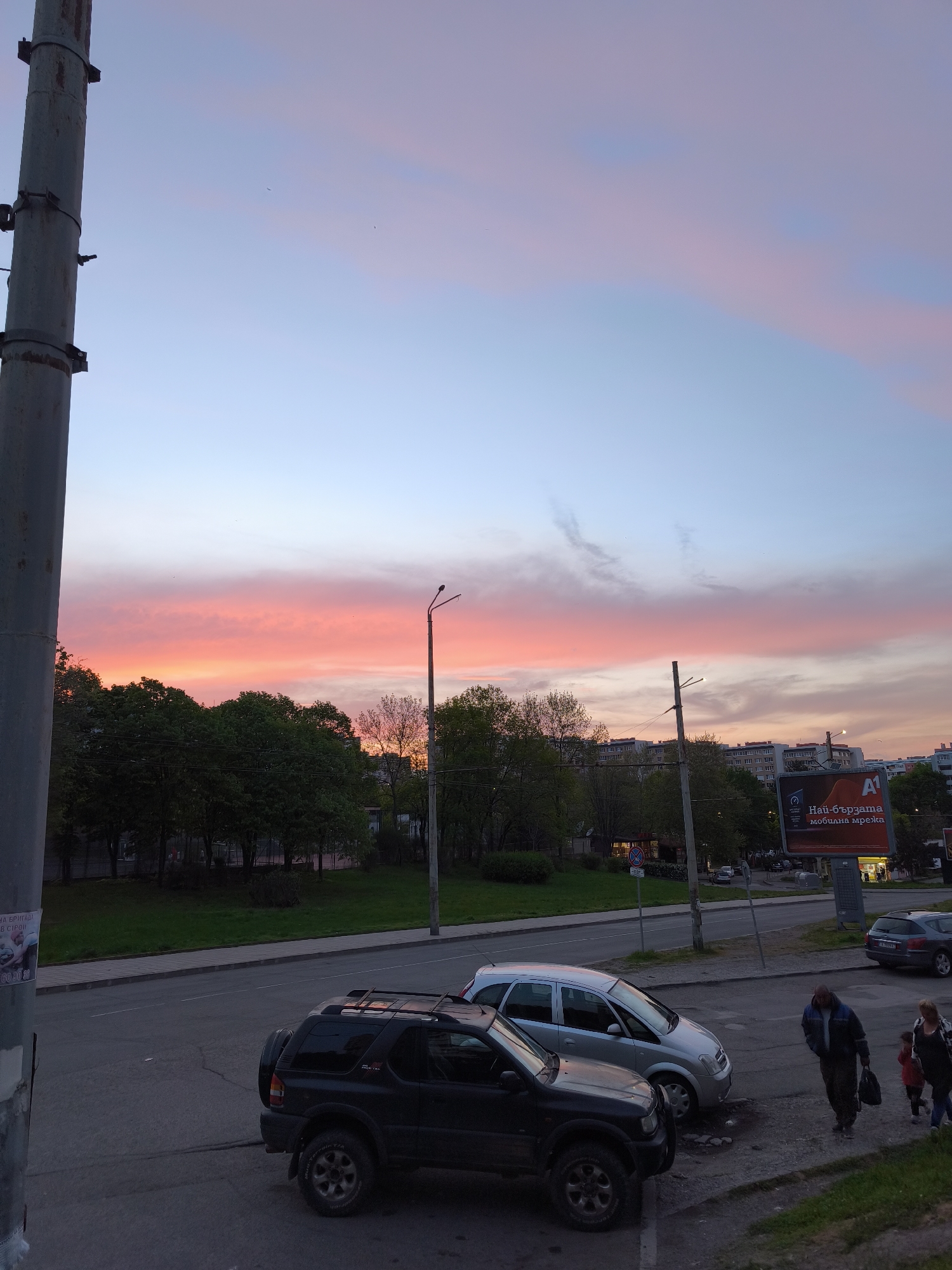 The image shows a street scene in Burgas, Bulgaria, at dusk. The sky is colored with shades of pink, orange, and blue. A row of trees separates the street from a green space. Two cars are parked along the curb: a dark-colored SUV and a silver hatchback. A man walks down the sidewalk carrying a bag, and a woman and child walk alongside him. A large billboard advertises A1 mobile services in Bulgarian. Utility poles are visible on the left and right of the frame. The overall lighting is dim. Buildings are visible in the background.