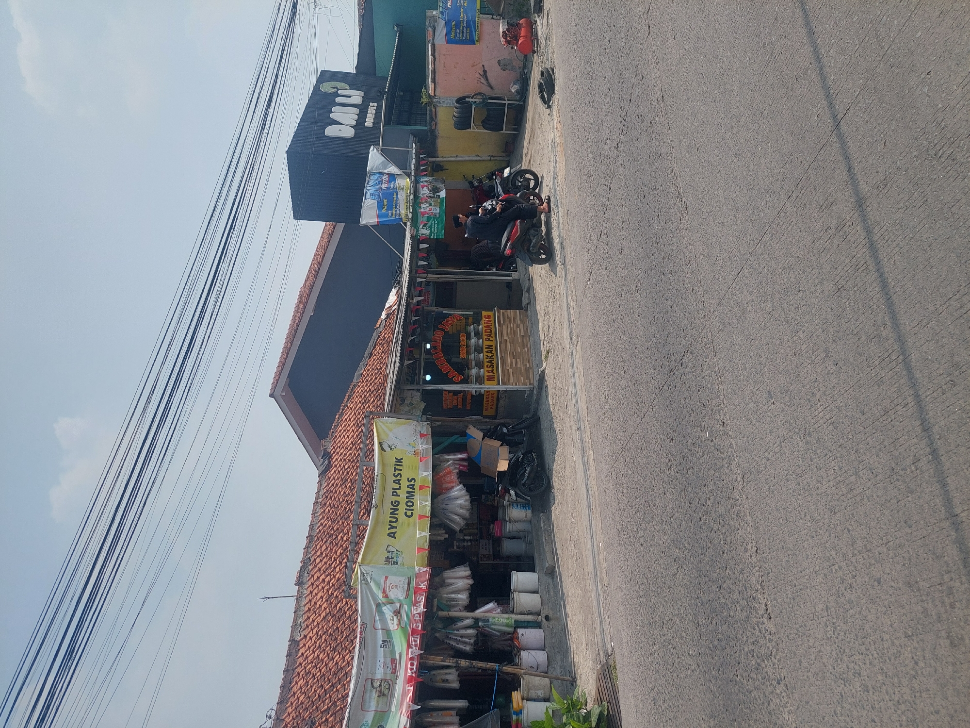The image depicts a street scene in Ciomas, Indonesia, under a clear sky. In the foreground, a paved road with some texture and cracks is visible, with a shadow extending from the right. The midground showcases a row of buildings and shops along the roadside. One shop, labeled "AYUNG PLASTIK CIOMAS," displays various plastic goods hanging from its facade and buckets on the ground. Adjacent to it, another establishment appears to be a food vendor, with signage indicating "MASAKAN PADANG" (Padang cuisine). A black motorcycle is parked in front of the plastic shop, and another motorcycle with two men on it is in motion in front of the food establishment. Further down the street, a dark-colored building with a prominent sign reading "DAILI" and "DONUTS" is visible. To the right of the "DAILI" building, another shopfront with signage in Indonesian and a red air compressor and tires suggest an automotive service. Overhead, a network of black electrical wires crisscrosses the sky. The overall environment suggests a commercial area in an Indonesian town or city.