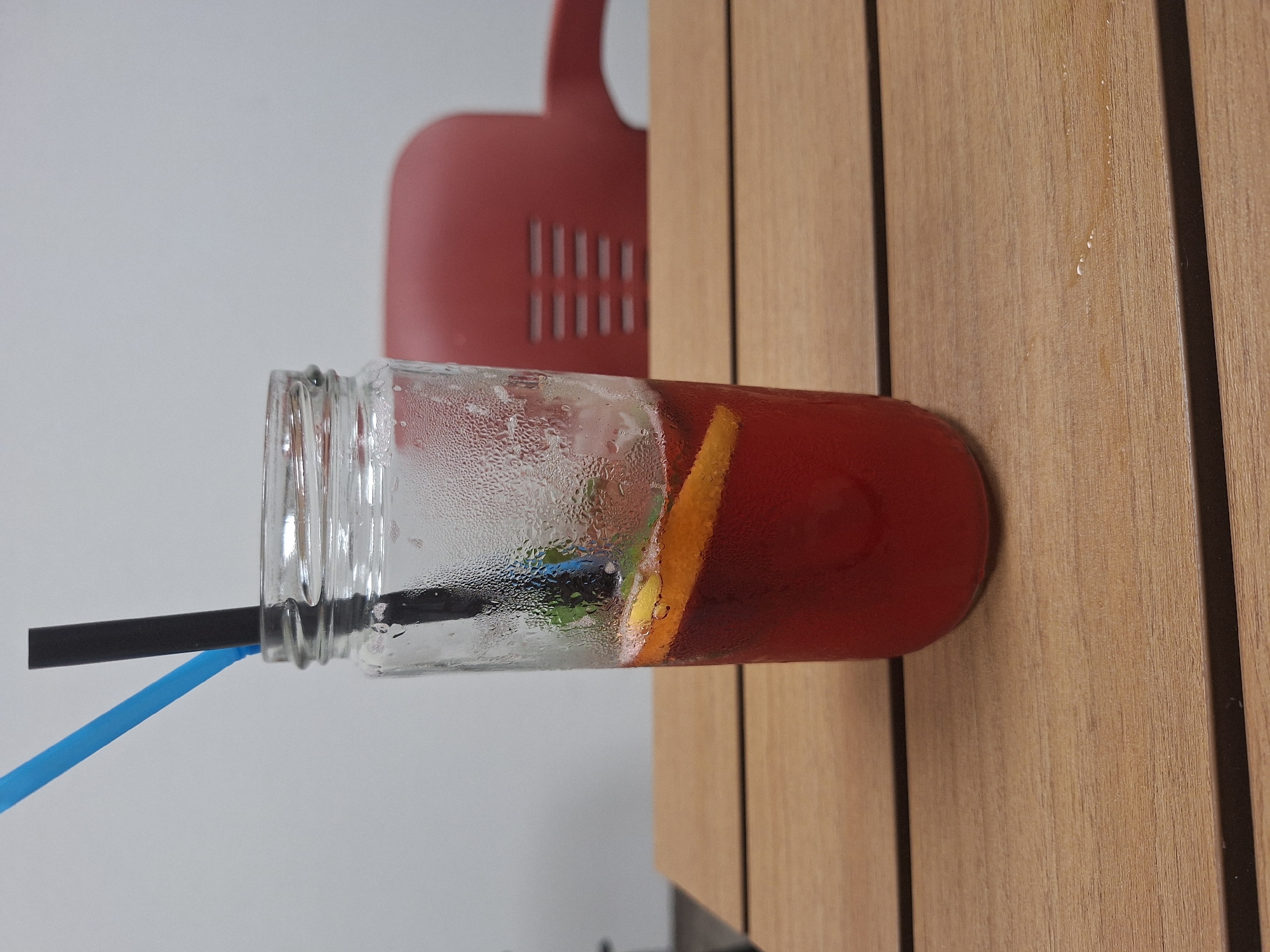 A refreshing, cold beverage served in a clear glass mason jar is the central subject of the image. The drink is a deep red color, likely a fruit punch or sangria, and contains slices of orange or other citrus fruit, along with subtle hints of green leaves, possibly mint. The glass jar is visibly covered in condensation, indicating the beverage is chilled.
Two straws are inserted into the drink: one standard black straw and one bright blue straw. The jar rests on a light-colored wooden surface, which appears to be composed of vertical planks, suggesting either a table or wall paneling.
In the blurred background, a prominent red object with a distinct curved shape and a pattern of vertical slits is visible. This object is set against a plain light gray or white wall. The scene suggests a casual setting, possibly indoors at a cafe, restaurant, or home. There are no people visible, and no text is present in the image. The image itself is originally oriented horizontally, rotated 90 degrees clockwise. The implied location context is Kraishte, Bulgaria.