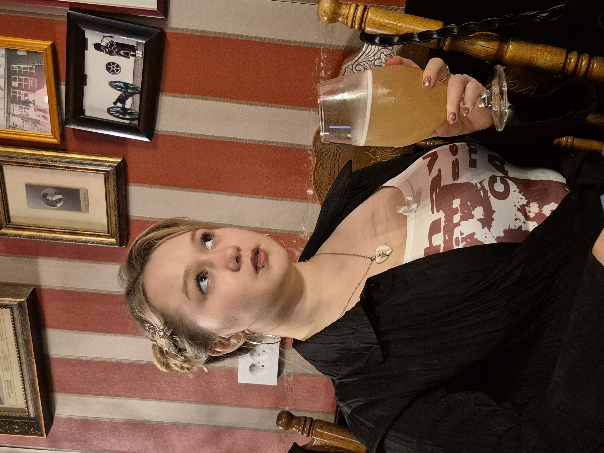 This media file shows a young woman with blonde hair pulled back in a bun, wearing a black, flowing top over a white shirt with a red, splotchy design. She has a heart-shaped pendant on a necklace and is wearing hoop earrings. Her fingernails are decorated with glitter. She is holding a glass of light-colored, frothy liquid, possibly beer. Her expression suggests a slight tilt of her head and an upward gaze. Behind her, the wall is decorated with horizontal red and beige stripes. There are several framed pictures on the wall. One frame on the left, with a gold finish, contains what appears to be a historical document or print. Above it is a smaller, dark-framed picture of an old building. To the right of that is another dark-framed picture, showing a black carriage with two large wheels and a person in uniform. A wooden chair with ornate carvings is visible to the right of the woman. The lighting suggests an indoor setting, likely in the evening or a dimly lit room. There is no visible text in the image. The scene appears to be a casual gathering or a visit to a bar or restaurant. The woman's expression is neutral.