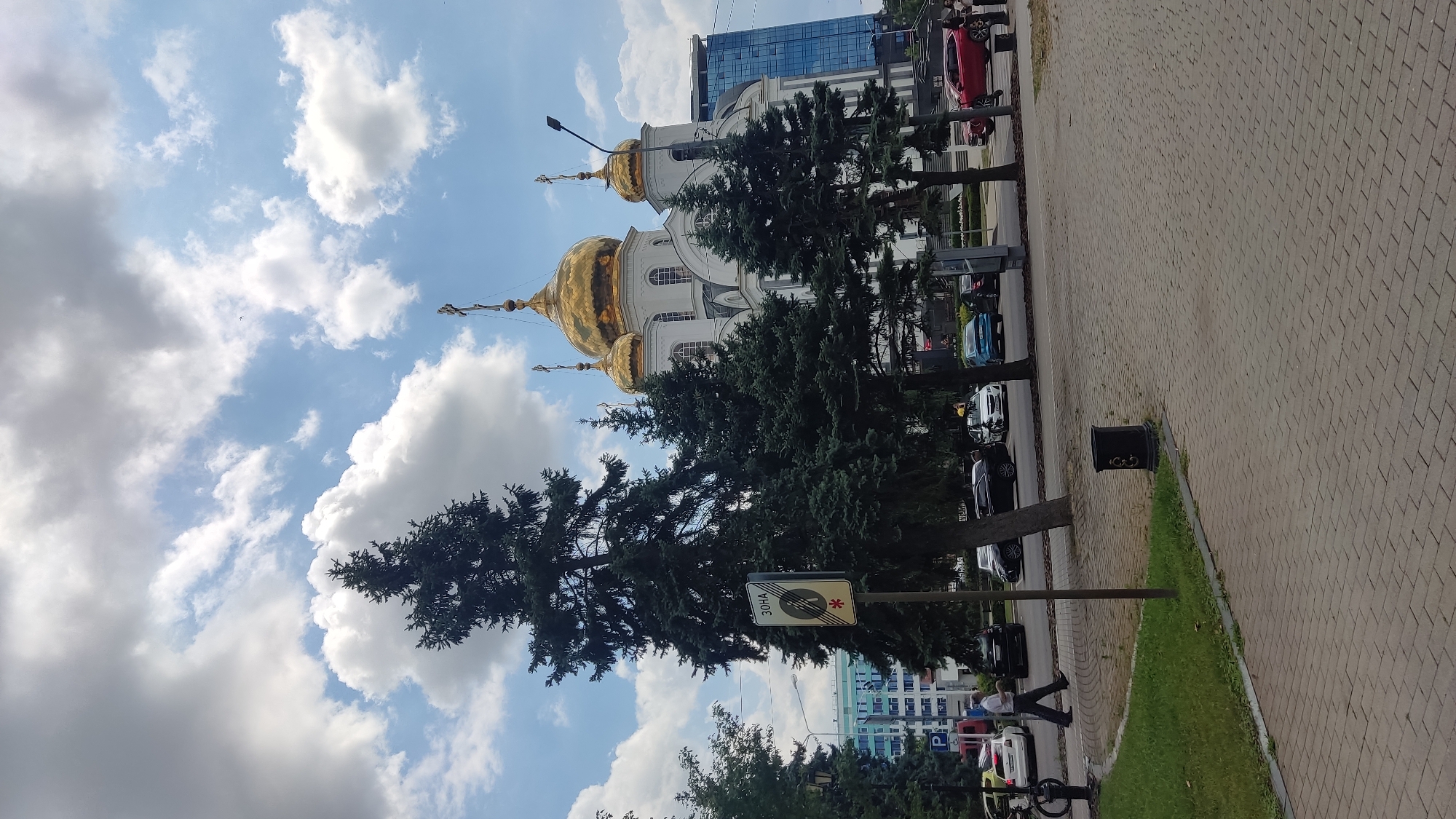This is a full-shot, eye-level photograph of the St. Vladimir Cathedral in Krasnodar, Russia. The cathedral is a prominent white building with golden domes, situated in the background. In the foreground, a paved walkway leads to a grassy area with several tall, dark green coniferous trees. A street with a few cars and a pedestrian is visible behind the trees. The sky is blue with scattered white clouds. A traffic sign with the Cyrillic word "ЗОНА" (zone) and a symbol of a car with a red cross over it is located to the left of the frame, near the trees. The overall scene depicts a sunny day in an urban setting, with a focus on the cathedral and its surrounding greenery.