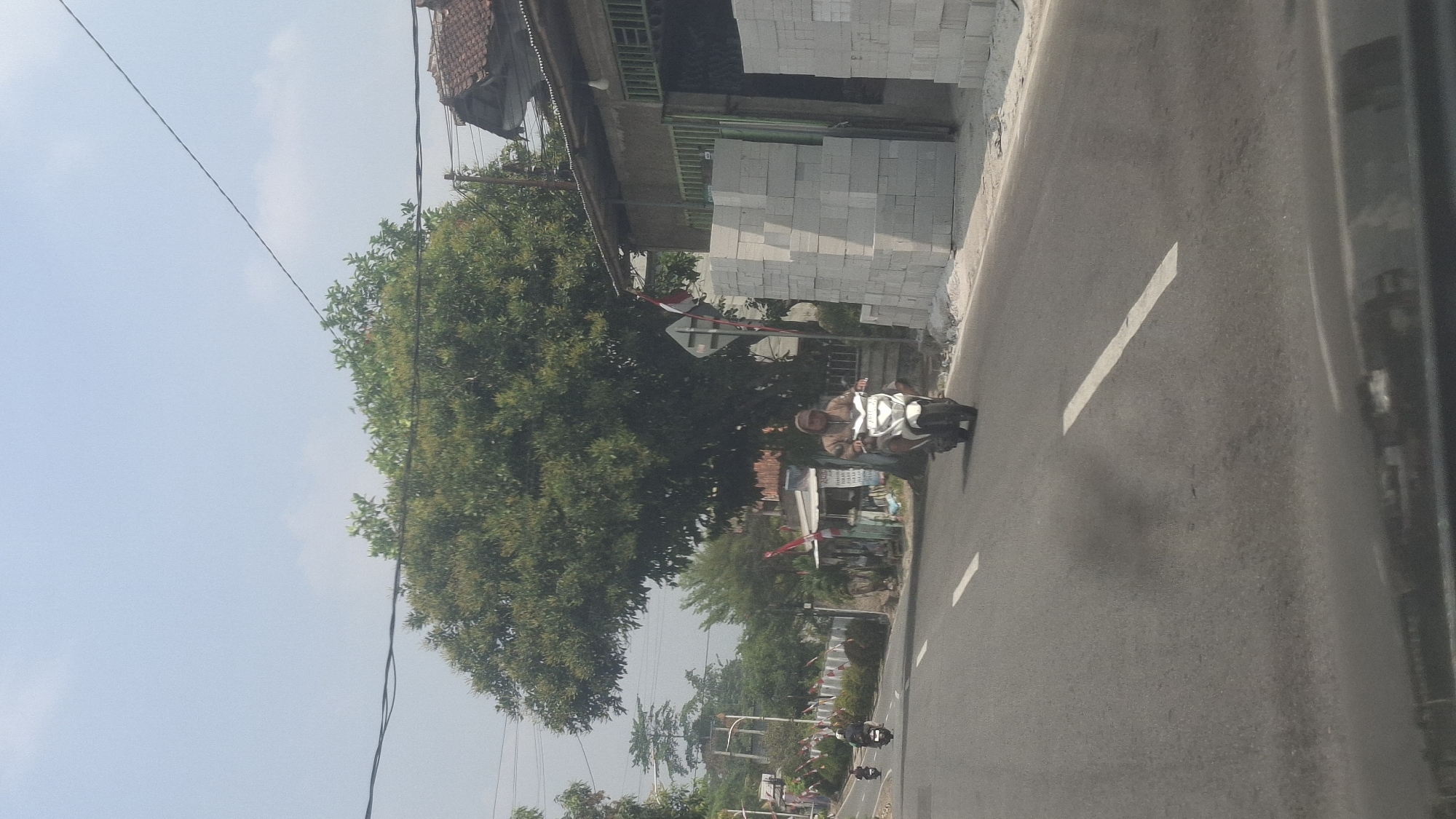 A light-colored motorcycle with a rider wearing a brown hoodie and a face covering is traveling down a paved road in Cilaku, Indonesia. The rider is in the foreground, positioned slightly to the right of the center of the image, heading towards the viewer. The road has white lane markings. To the right of the road, a pile of light-colored building blocks is stacked against the side of a building with a corrugated metal roof. A large tree with dense green foliage is behind the motorcycle, and a street sign with red and white flags is visible near the tree. In the background, a few other motorcycles are seen further down the road. The sky is clear and light blue.
