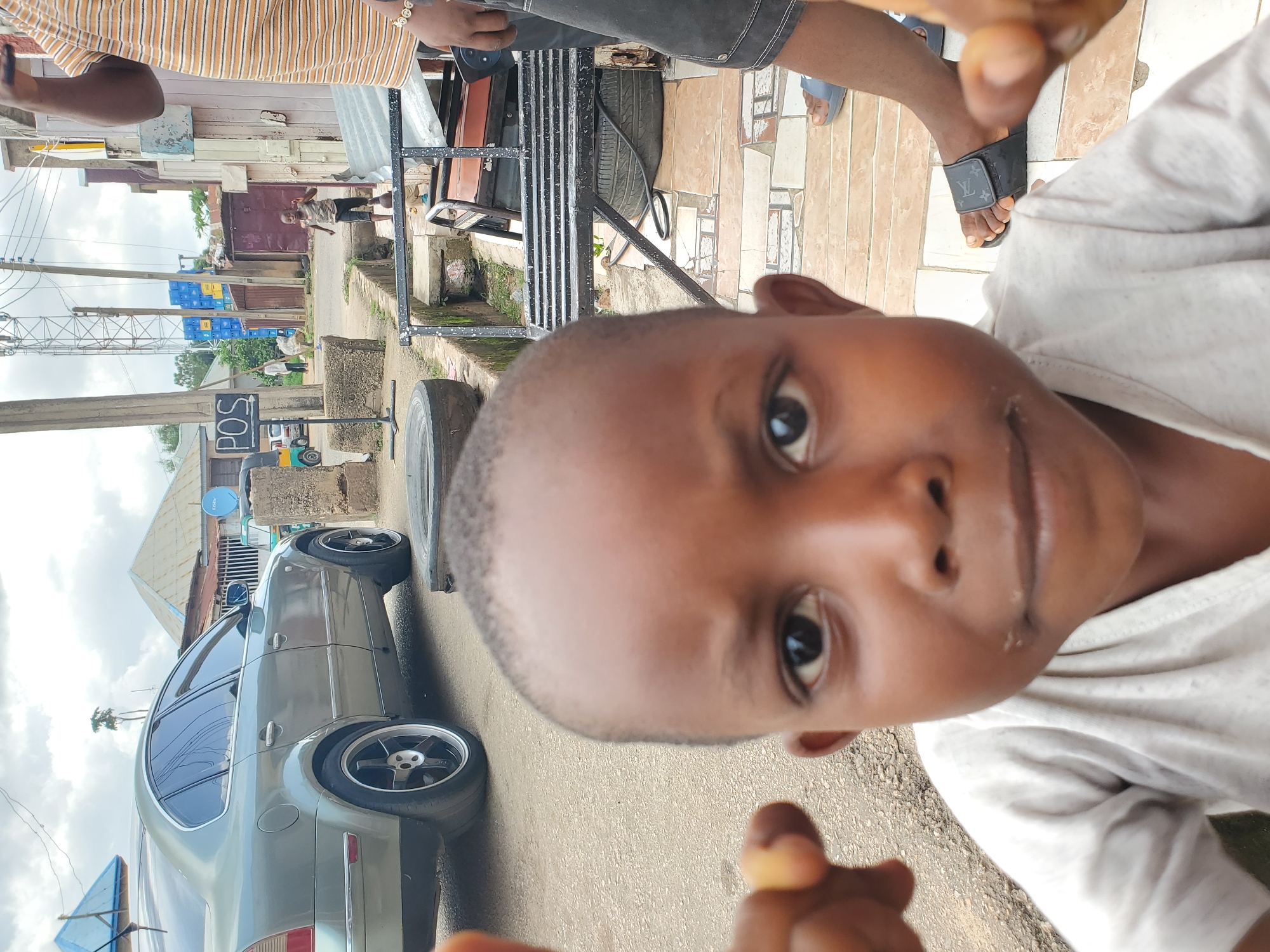 This media file shows a close-up, low-angle shot of a young boy's face, filling most of the frame. His eyes are looking directly at the camera, and his mouth is slightly open. He appears to be wearing a light-colored t-shirt. In the background, the image is oriented vertically and shows a street scene in Jos, Nigeria. A silver sedan is parked on the right side of the road. Further down the street, there are buildings and stacks of blue crates. The sky appears cloudy, suggesting it might be an overcast day. The overall mood seems informal and candid, possibly indicating a moment captured spontaneously. No visible text is clearly discernible in the image.