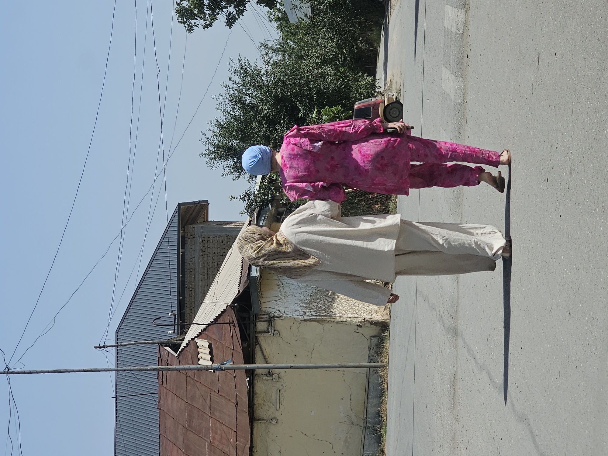 Two women are walking away from the camera on an asphalt road in Özgön, Kyrgyzstan. The woman on the left is wearing a light-colored headscarf and a loose, long-sleeved top and pants. The woman on the right is wearing a bright blue headscarf and a vibrant pink floral-patterned outfit, also a long-sleeved top and pants. They appear to be walking side-by-side, with the woman on the left's left arm slightly extended towards the woman on the right. In the background, a one-story building with a red corrugated metal roof is visible on the left, adjacent to a lighter colored structure with a light-colored, possibly corrugated, roof. Trees and foliage line the right side of the road. A small, red car is parked on the right side of the road in the middle ground. Power lines are visible crisscrossing the sky above the scene. The sky is clear and blue. The ground is paved with asphalt, showing some cracks and unevenness.