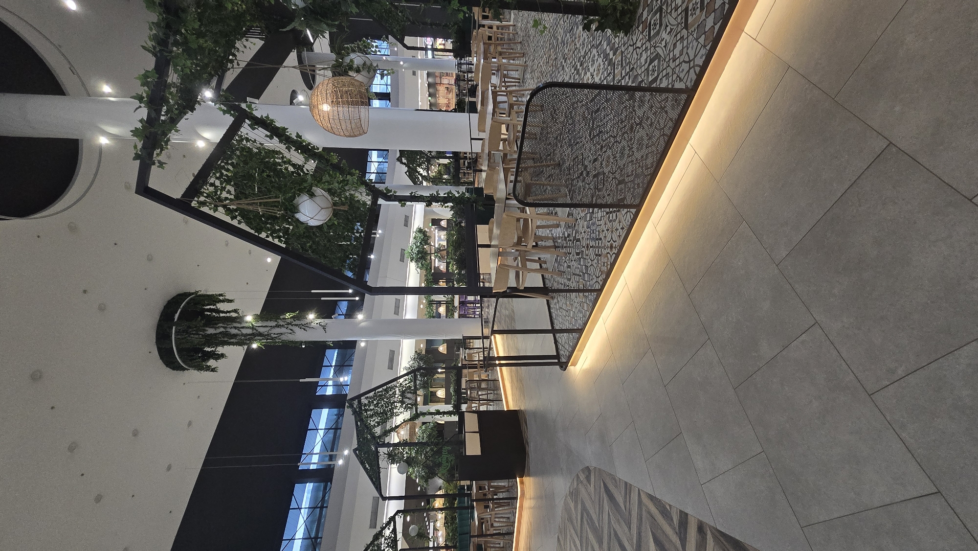 The image depicts an interior scene, likely a shopping mall or commercial space, in Stara Zagora, Bulgaria. The focal point is a restaurant area with rows of light-colored wooden chairs and tables. The flooring is patterned with decorative tiles near the dining area, transitioning to large gray stone tiles in the foreground. Overhead, there are black metal structures adorned with artificial greenery and hanging decorative lights. The ceiling is high with several recessed lights. Natural light filters through large windows or skylights. The space feels modern and designed. There are no people in the immediate view.