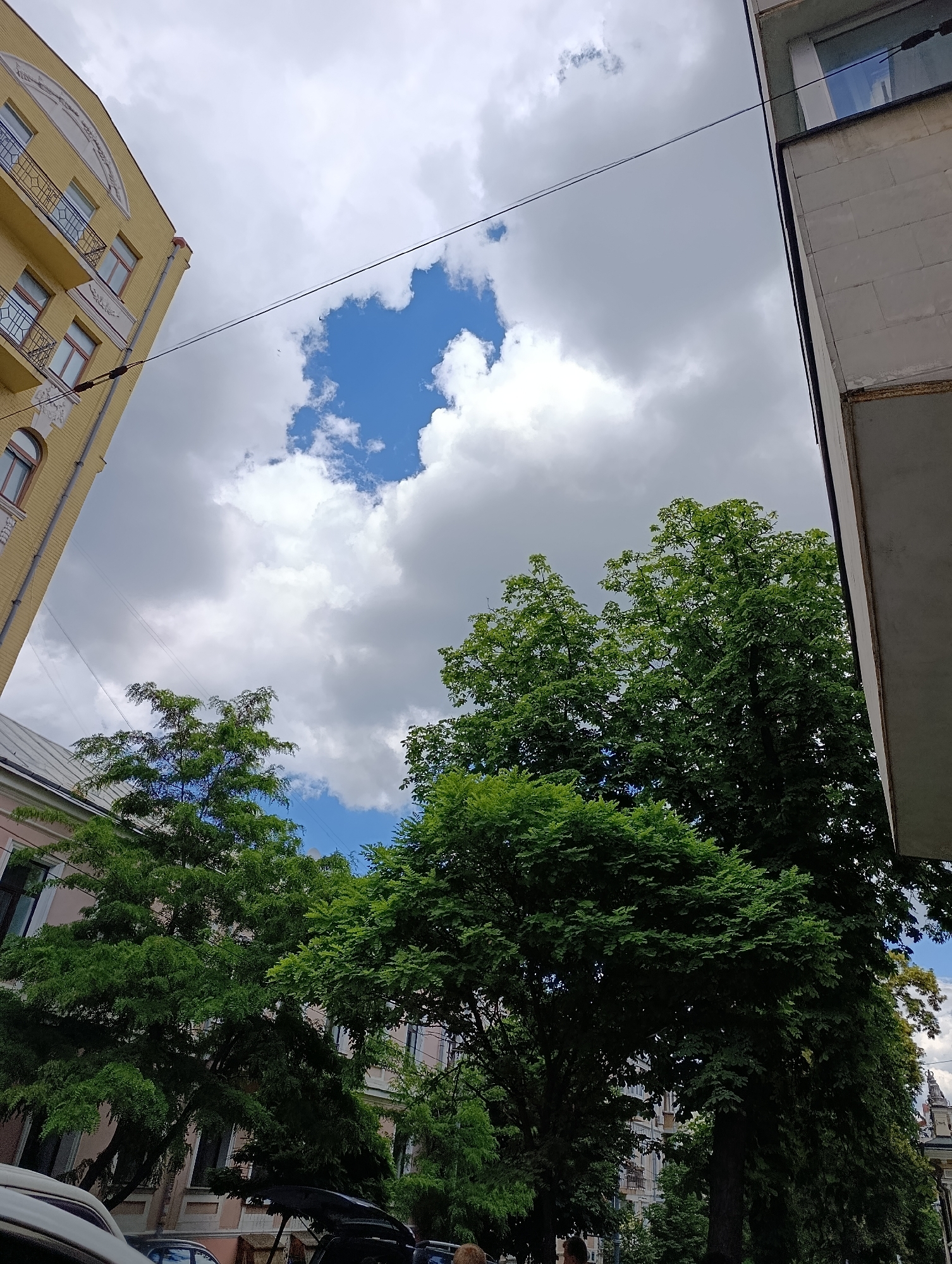 The image shows a view of buildings and trees under a cloudy sky, with a patch of blue sky visible. A yellow building with balconies is on the left, and a light-colored building is on the right. Lush green trees are in the foreground and midground. The scene is likely a street in Kyiv, Ukraine. The weather appears to be overcast with some sunny breaks. No people or specific actions are clearly discernible in the image.